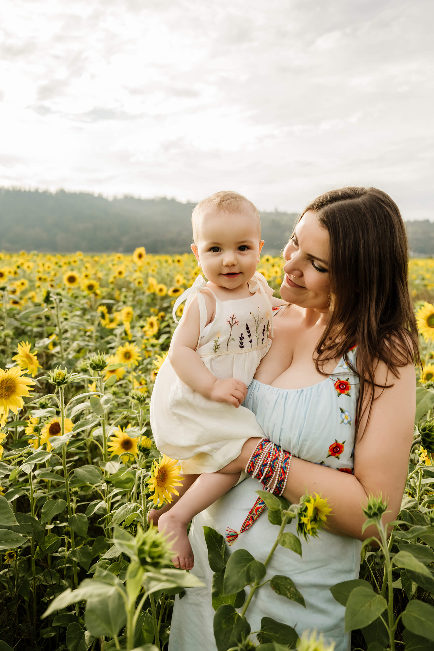 Mother holds her smiling baby among tall sunflowers, both bathed in warm evening light with wooded hills beyond.