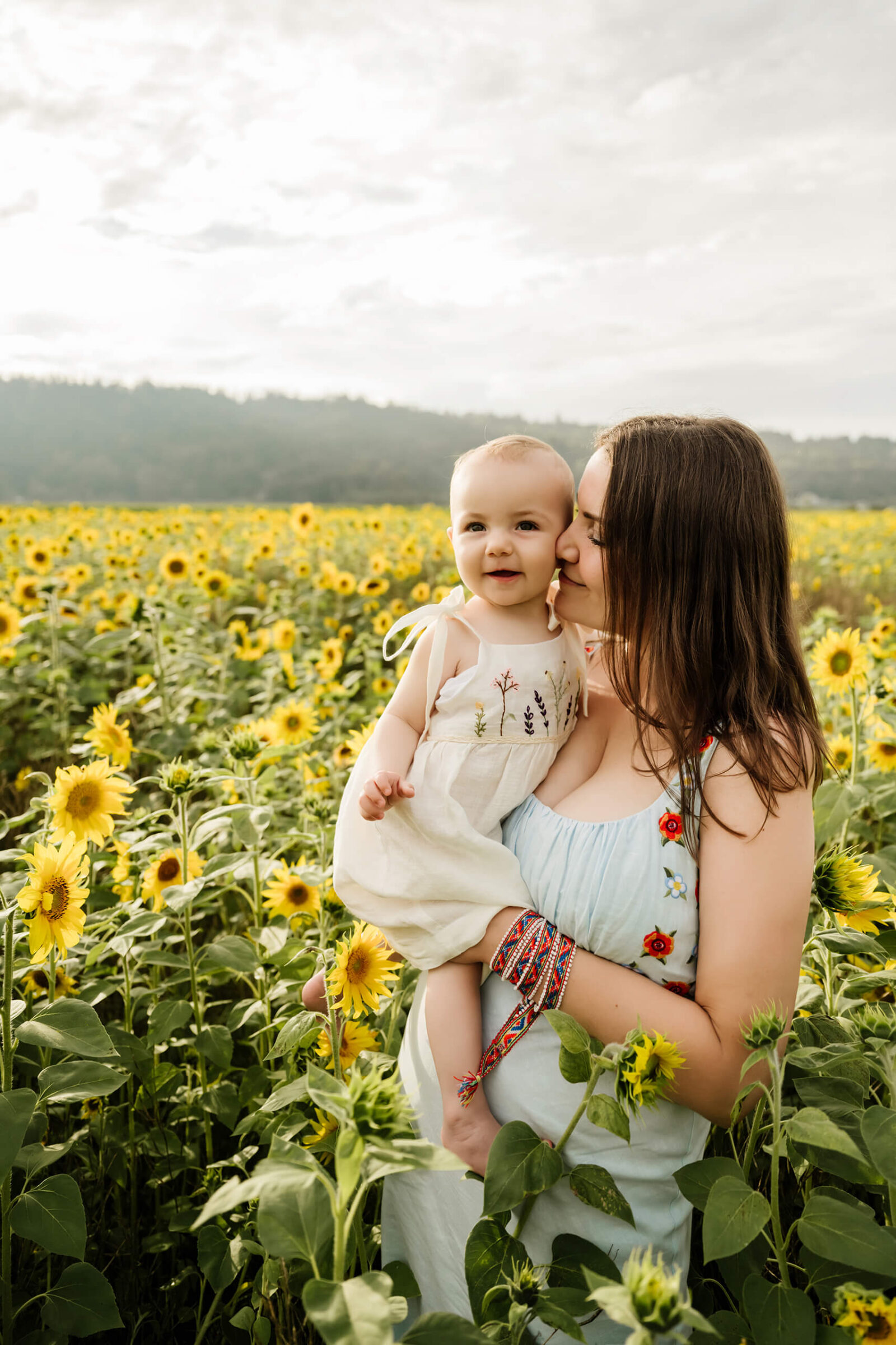 Mother kisses her smiling baby’s cheek while standing in a dense, sun-lit sunflower field.