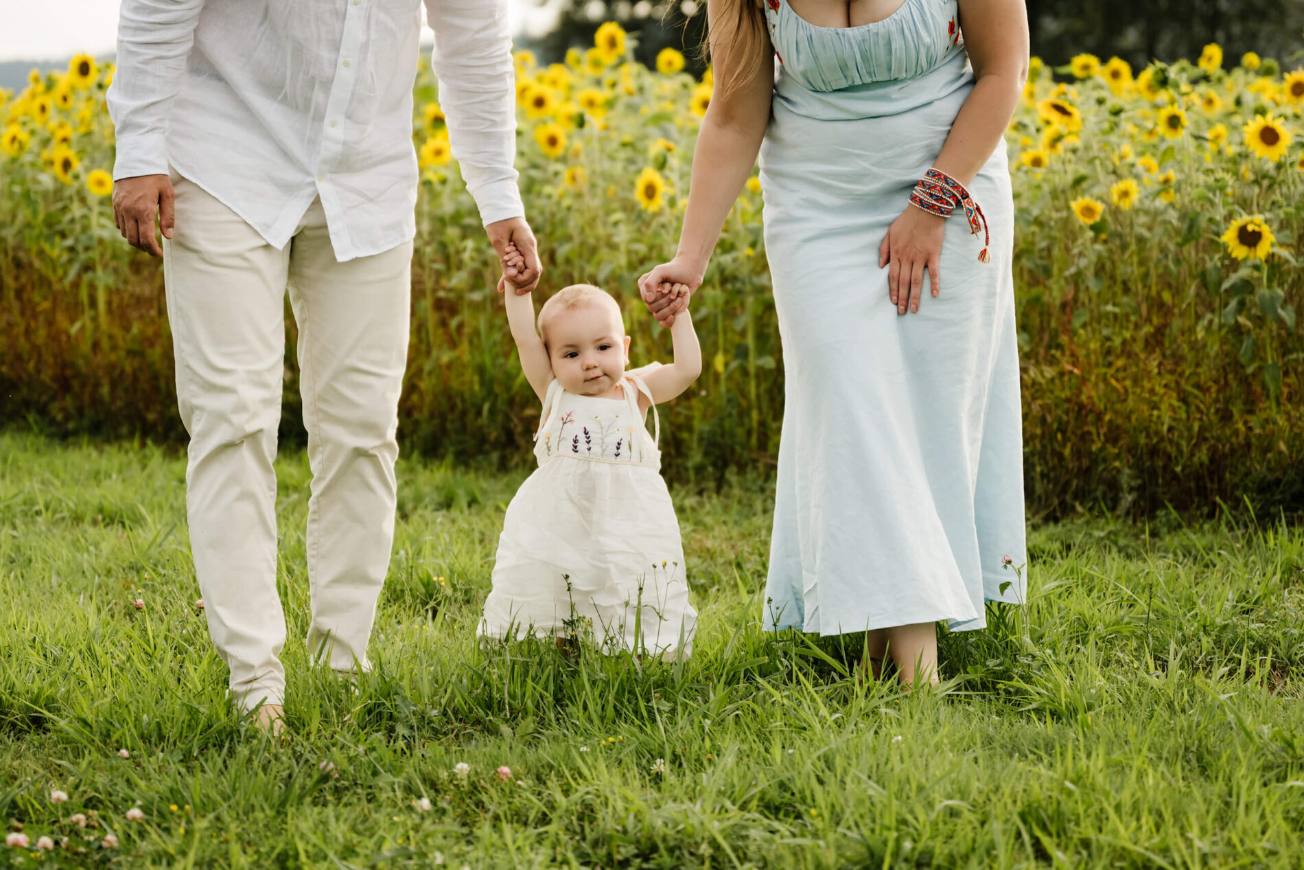 Cropped view of parents helping their toddler take steps on green grass, with yellow sunflowers behind them.