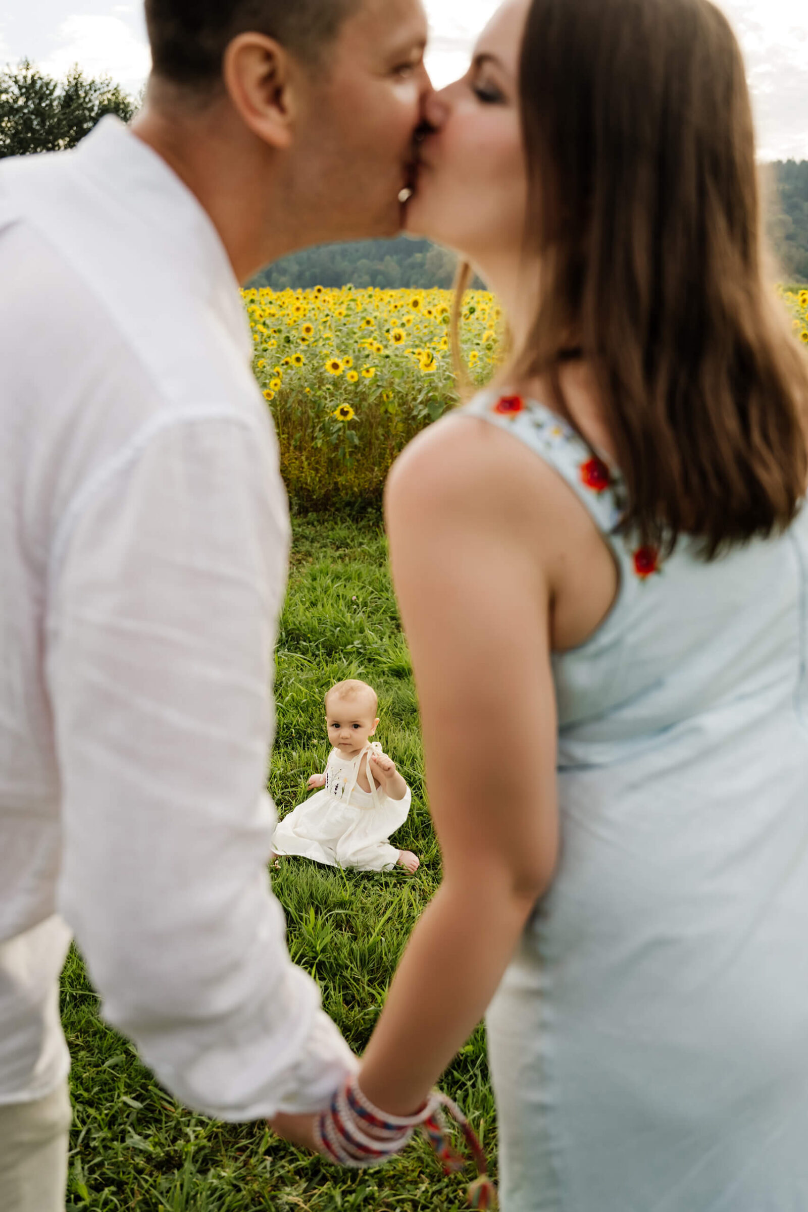 In-focus baby sits on grass between blurred parents sharing a kiss, framed by a bright sunflower backdrop.