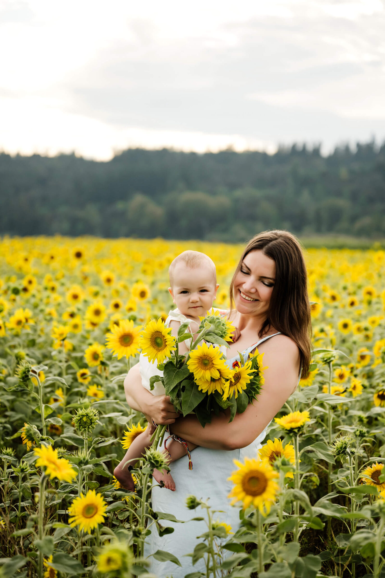 Mother holds baby and a freshly picked bouquet of sunflowers, both beaming amid endless blooms.