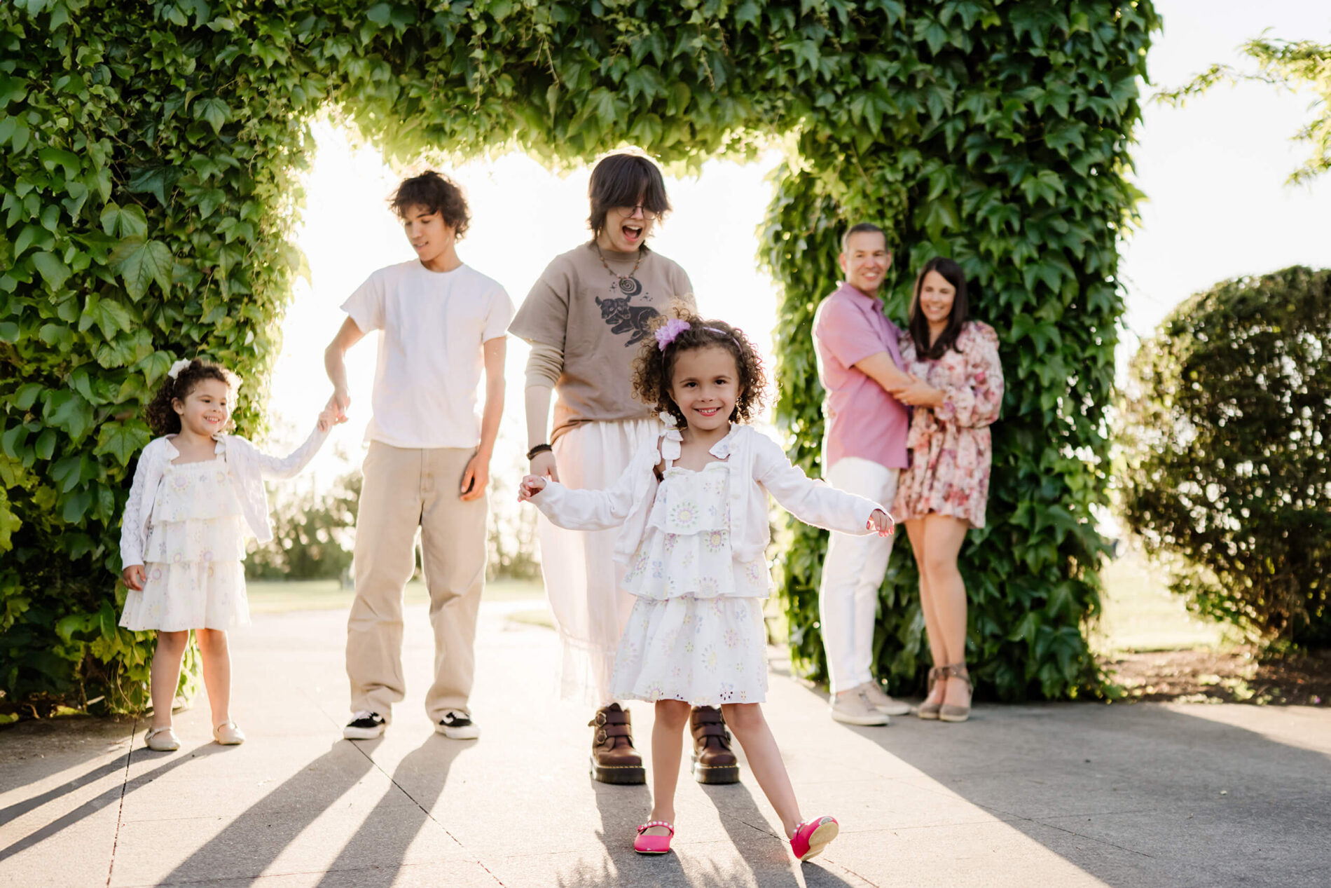 Little girl in a white sundress skips toward the camera while her family smiles beneath a sun-lit ivy archway at Heritage Park in Kirkland, WA.