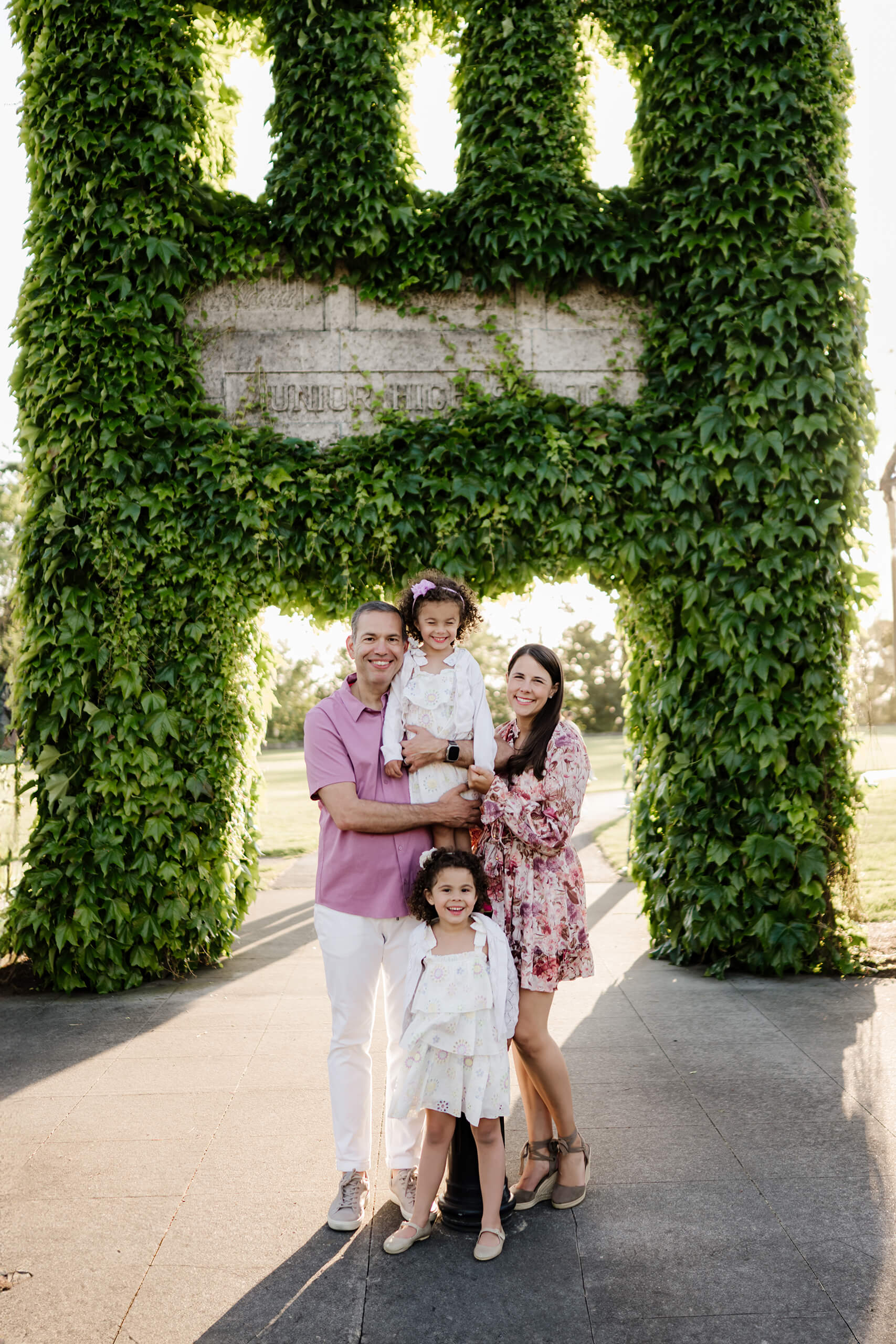 Parents hold their two young daughters below an ivy-draped archway glowing in late-afternoon light at Heritage Park in Kirkland, Washington
