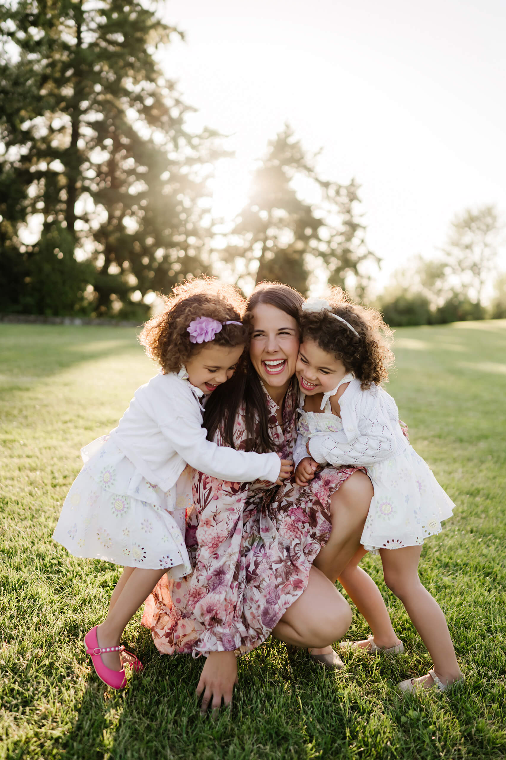 Mother kneels and laughs as her twin daughters hug her tightly on the sun-lit lawn of Heritage Park in Kirkland, WA.