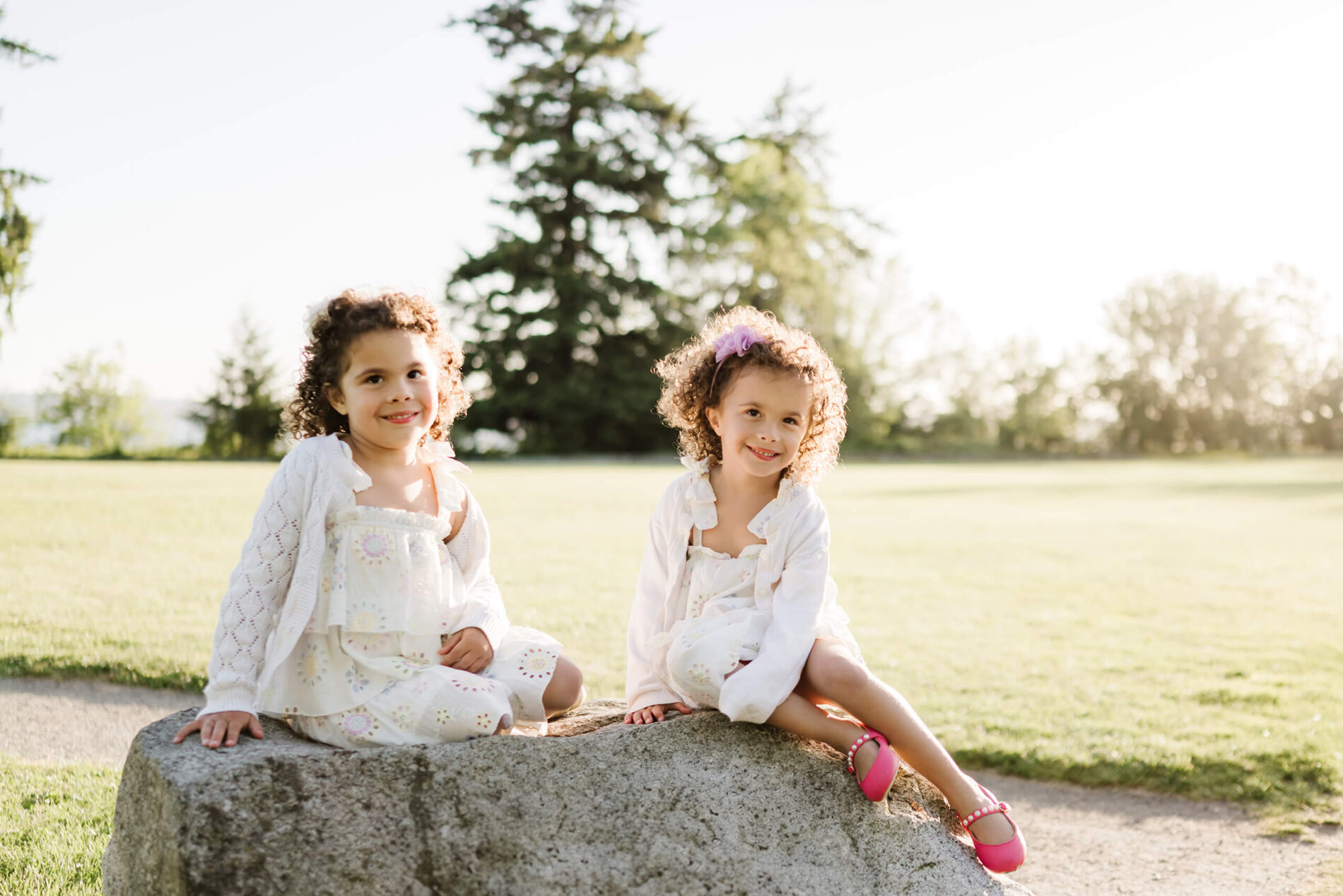 Two curly-haired sisters in matching eyelet dresses sit on a large rock in an open meadow at Heritage Park, Kirkland, WA.