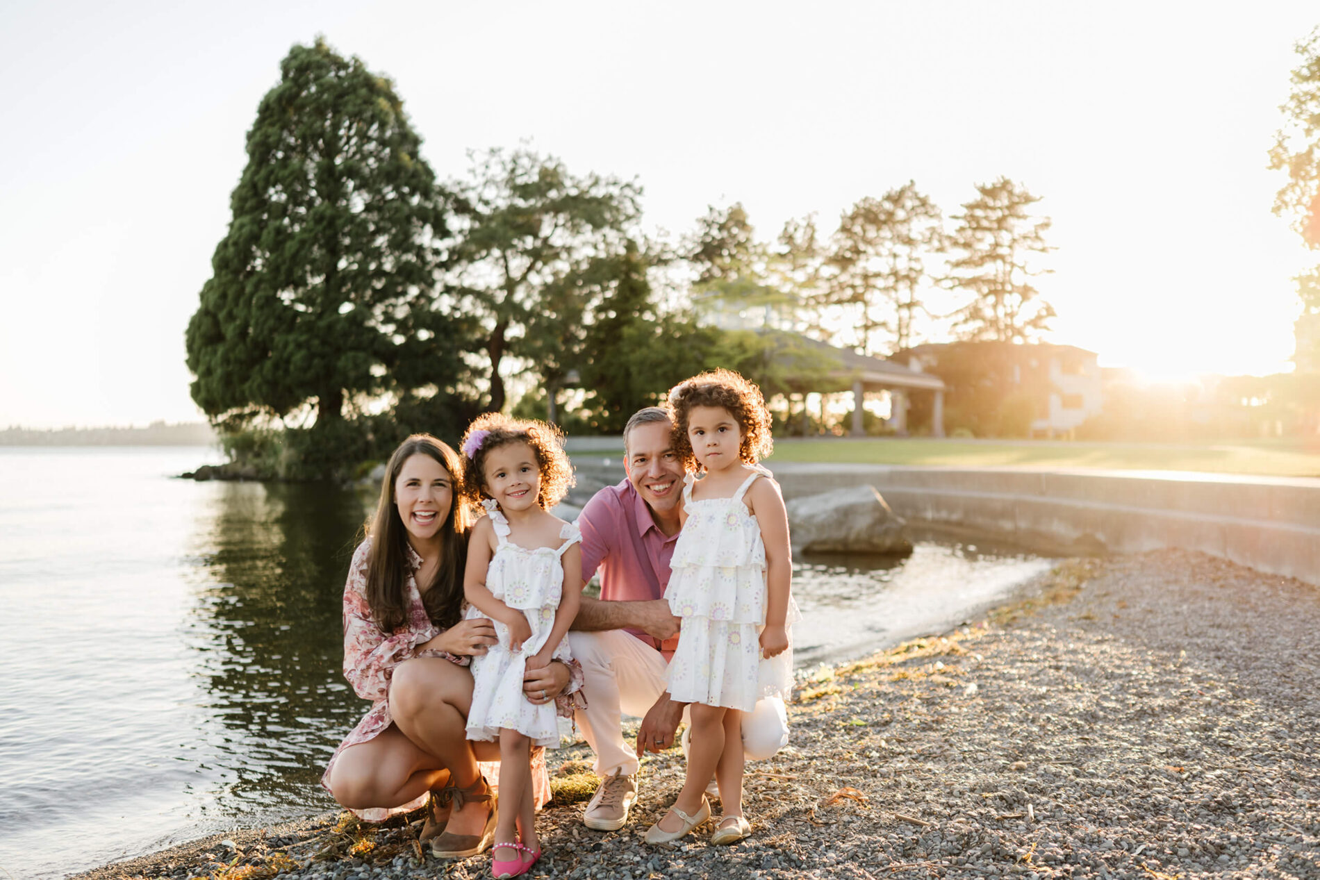 Parents kneel beside Lake Washington with their twin daughters, laughing in golden-hour light at Marina Park in Kirkland, WA.