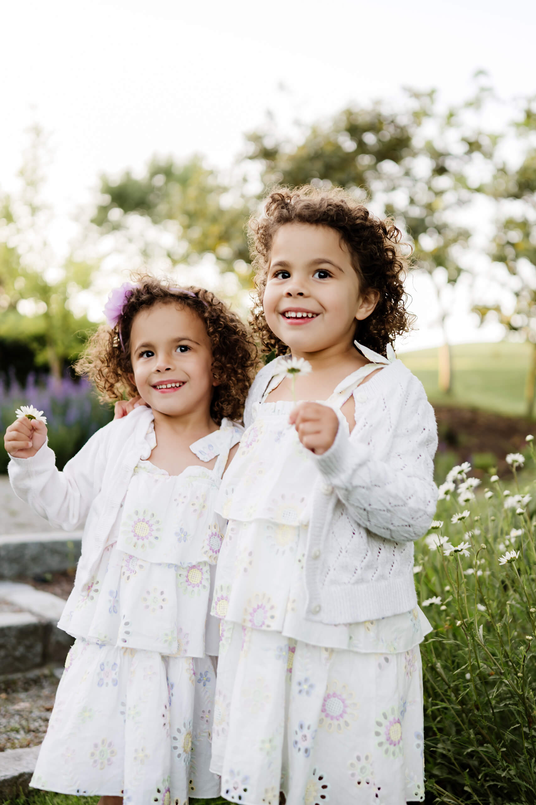 Twin curly-haired girls clutch daisies and smile amid garden blooms at Marina Park Public Beach, Kirkland, Washington.