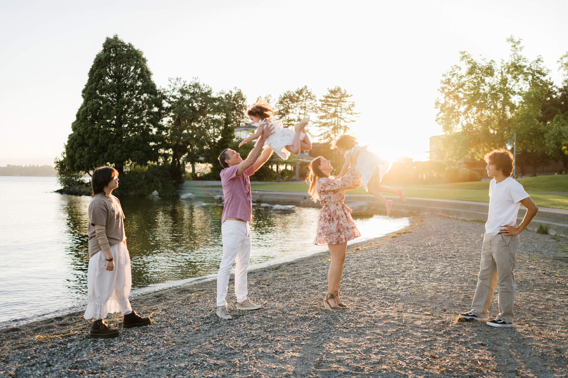 Mom and dad joyfully toss their young daughters into the air while siblings watch on the lakeside beach at Marina Park, Kirkland, WA.