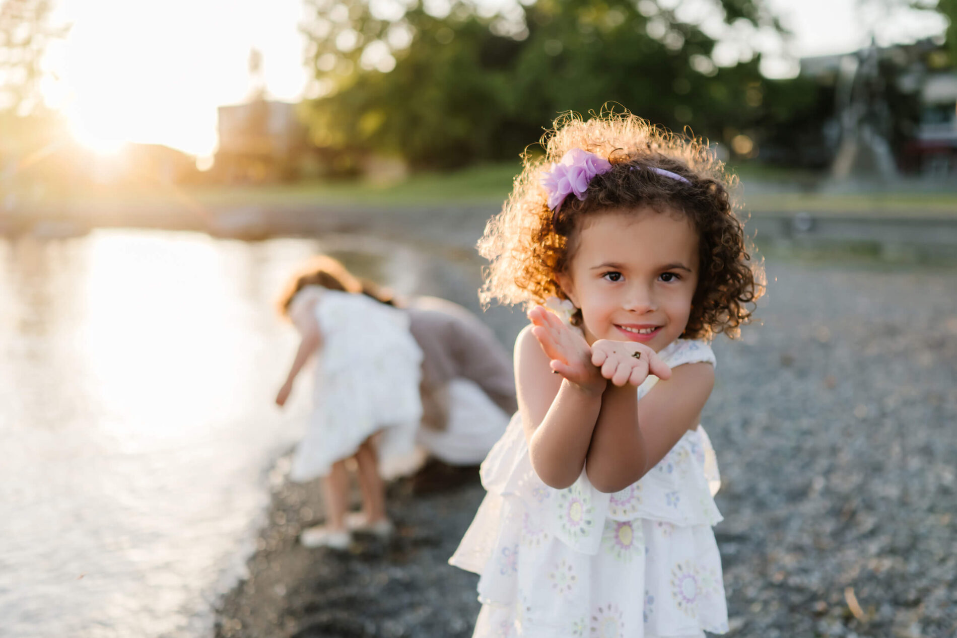 Little girl proudly shows pebbles cupped in her hands on the beach, lake sparkling behind her at Marina Park, Kirkland, WA.