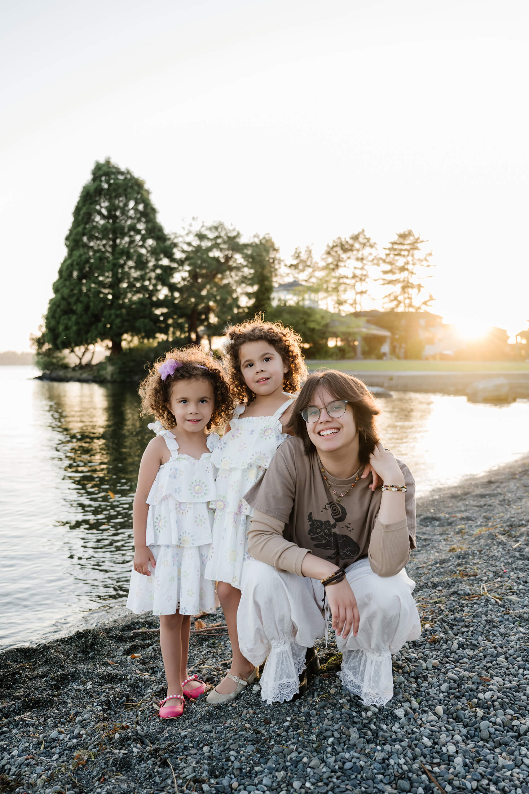 Teen sister crouches on the rocky lake shore with her twin siblings, all smiling at sunset in Marina Park, Kirkland, WA.