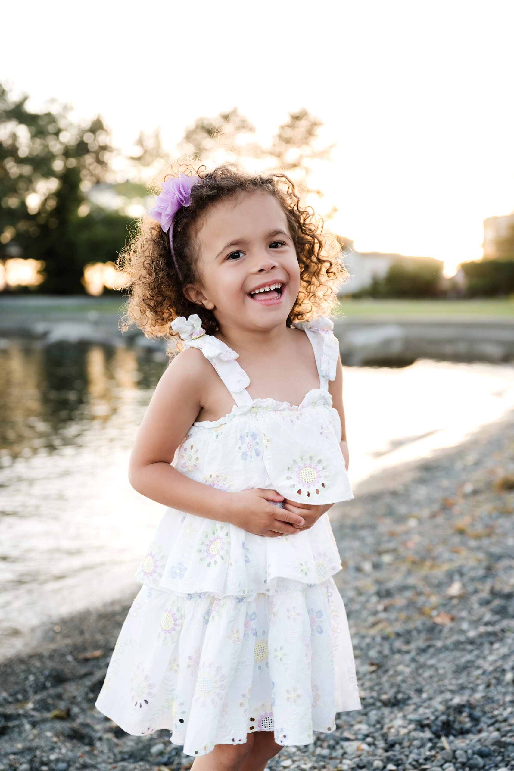 Close-up of a laughing curly-haired girl in a white sundress beside the water at Marina Park in Kirkland, WA.