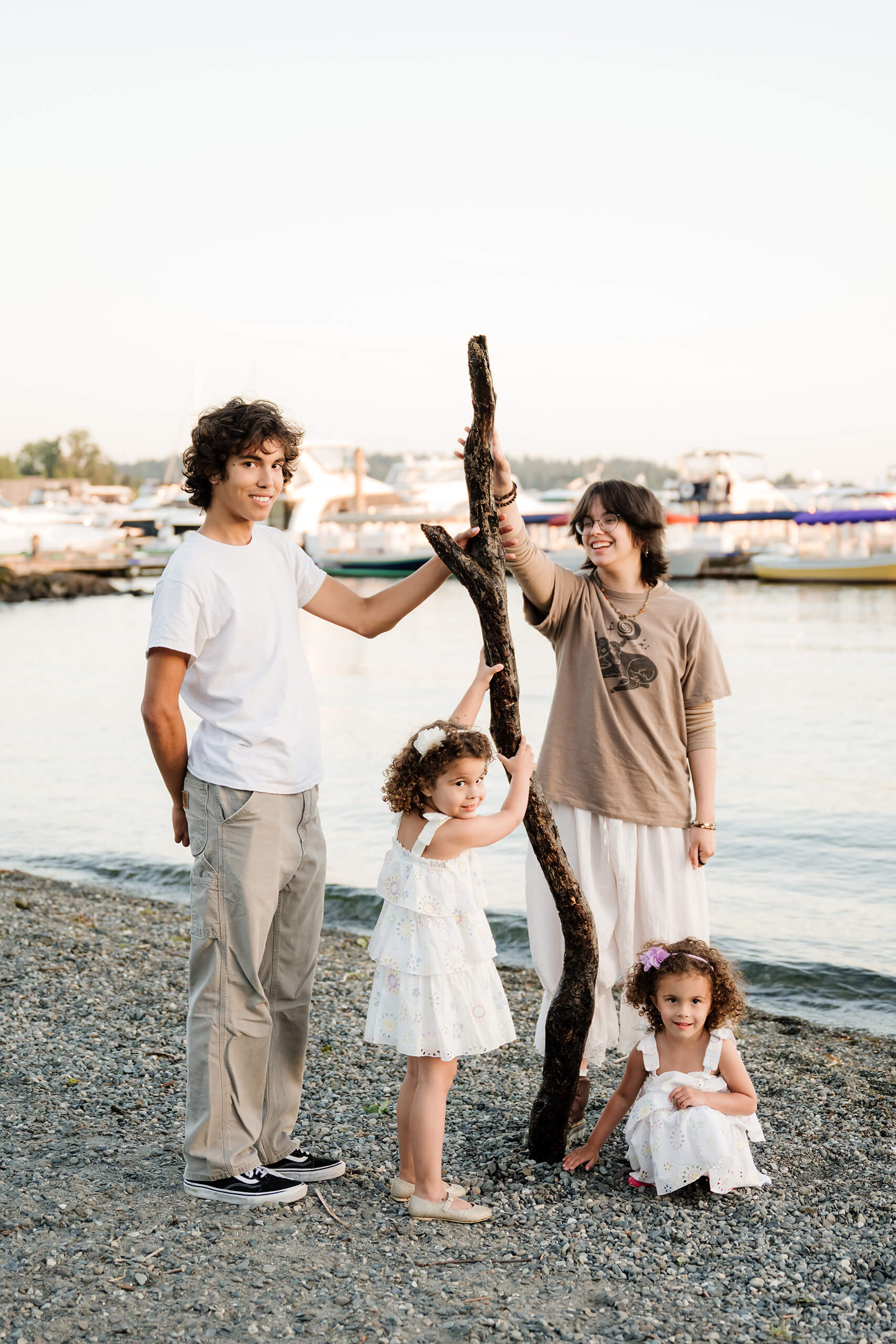 Older siblings help their little sisters balance a large driftwood branch on the marina’s pebble beach with boats in the background at Marina Park, Kirkland, WA.
