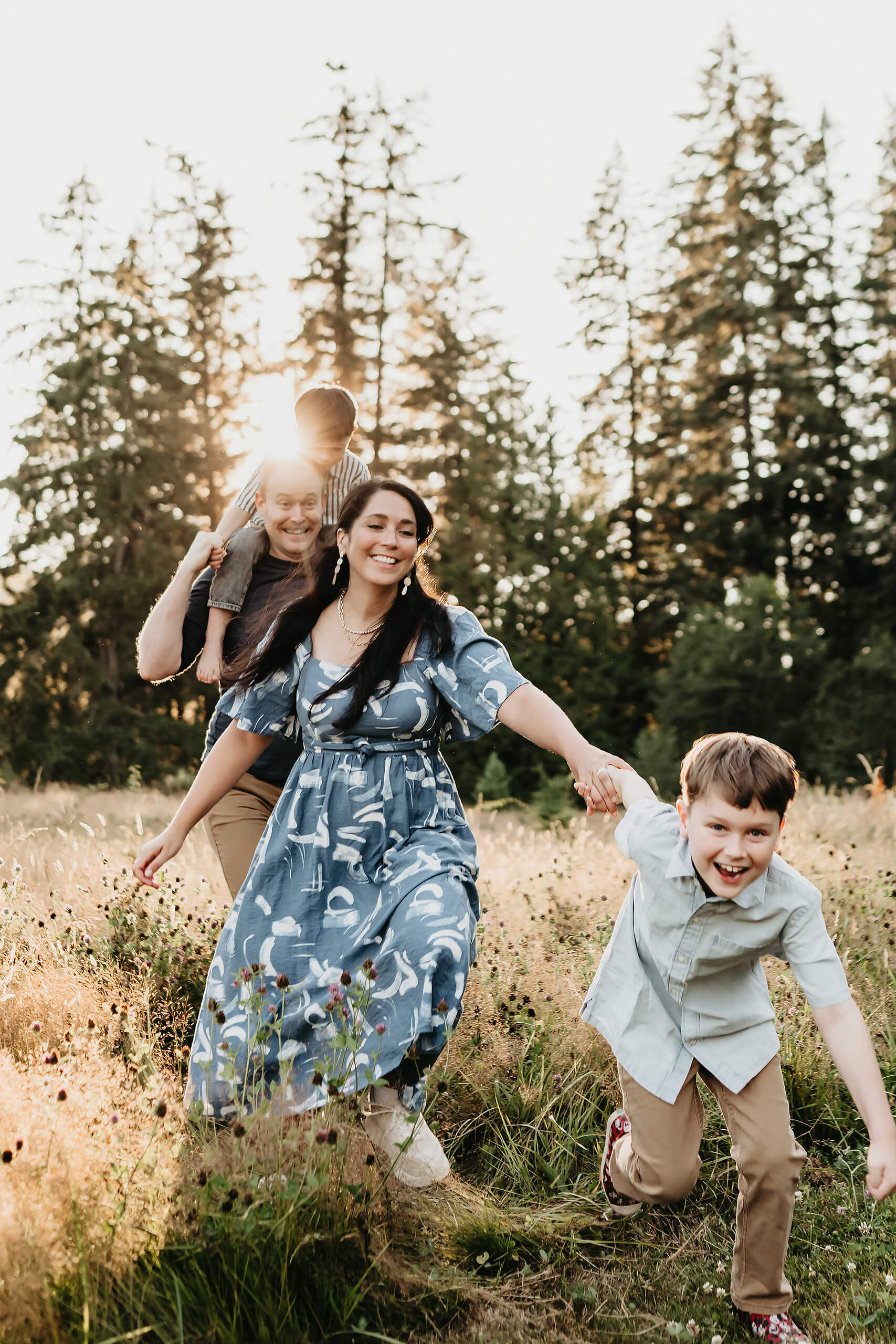 Family of four running through a sun-lit Redmond meadow; mom in a blue patterned dress leads laughing son while dad follows with child on his shoulders at golden hour.