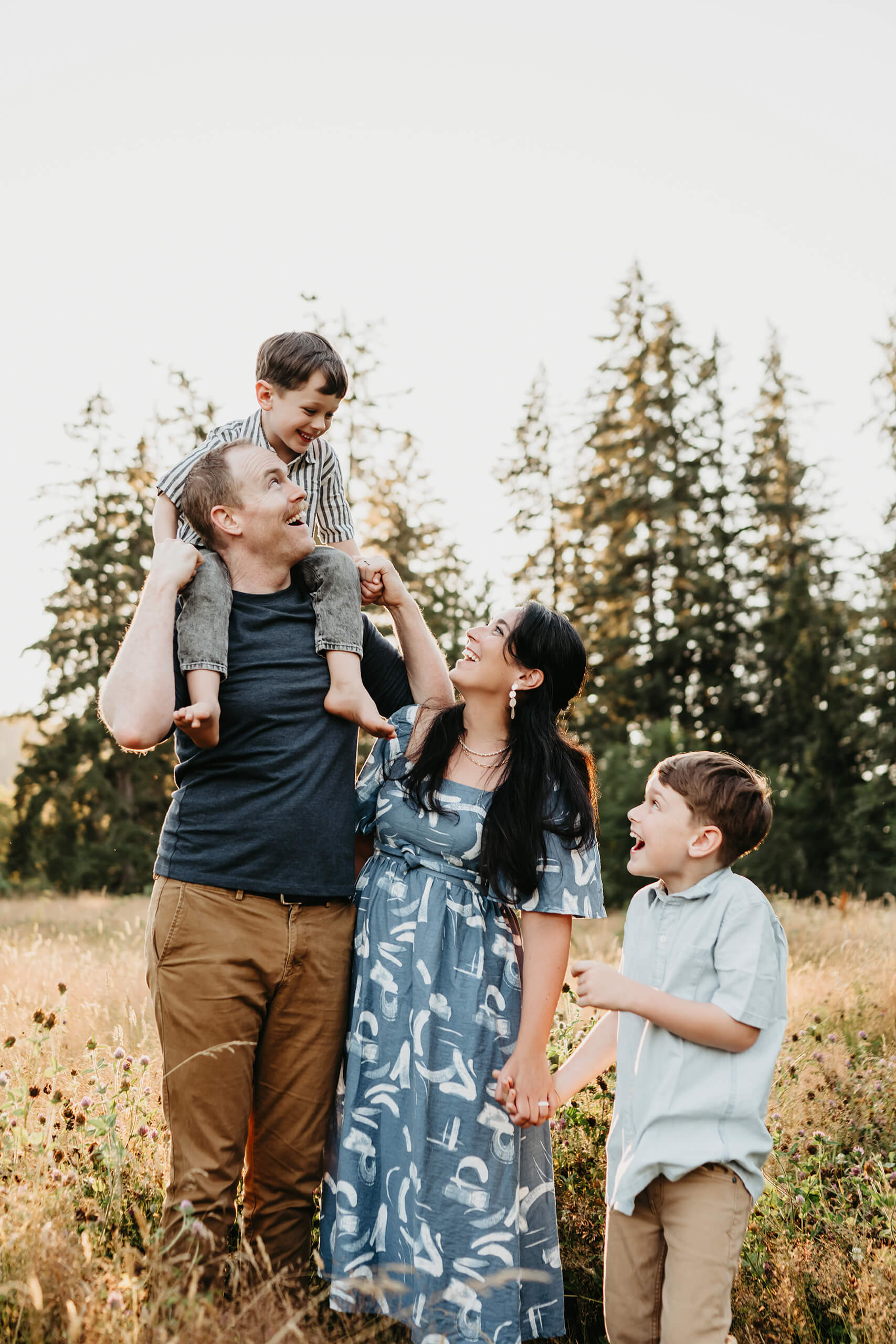 Playful Redmond family portrait at sunset: dad hoists son on shoulders, mom in blue dress laughs while holding younger boy’s hand amid tall grass and evergreens.