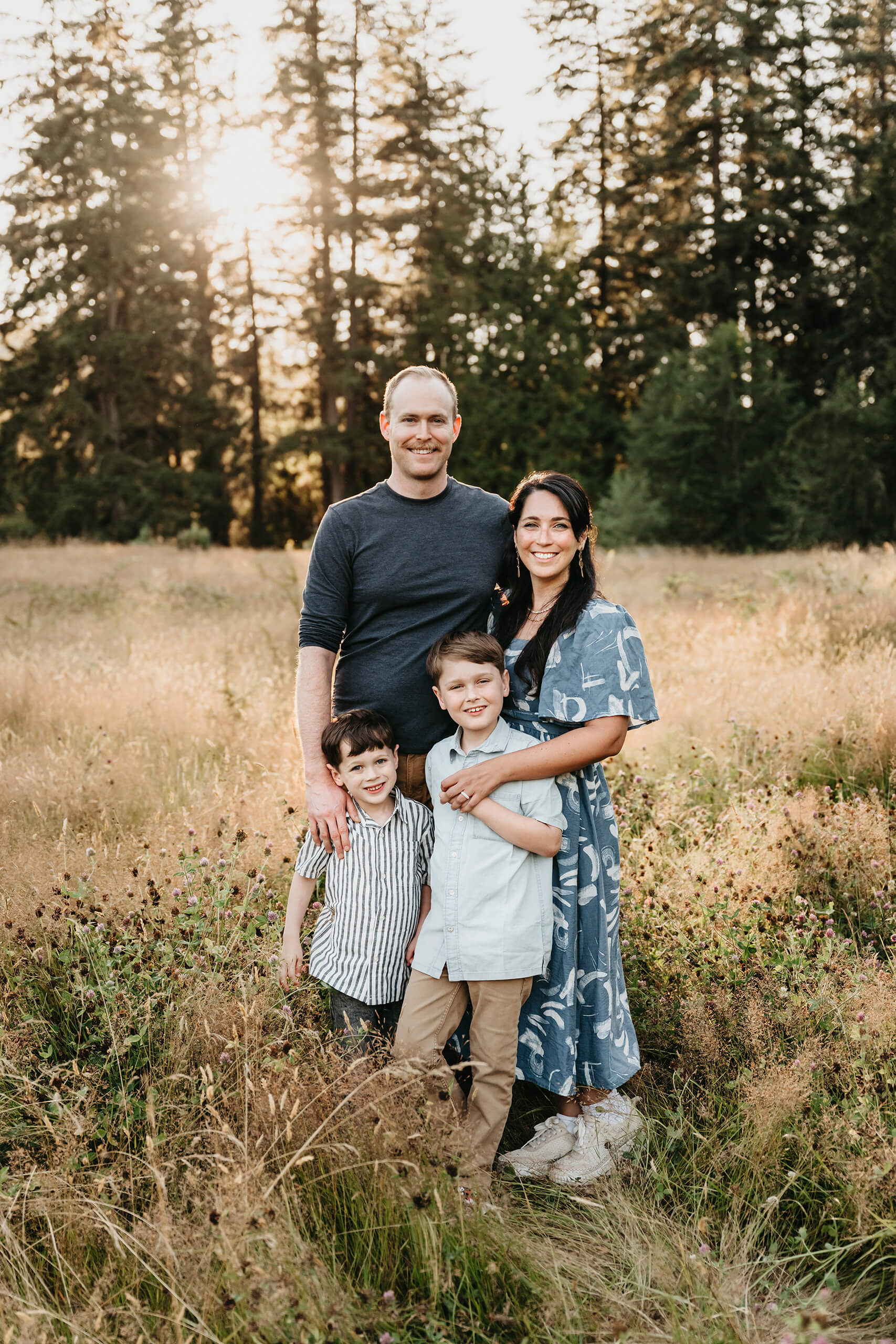 Family of four standing close in a sun-lit meadow, evergreen trees glowing behind them at sunset.