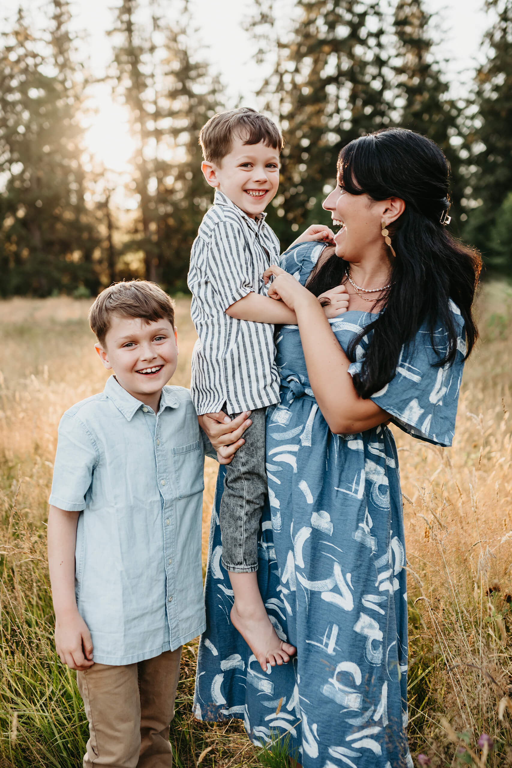 Mom laughing while holding one son in her arms as the other stands beside her, all bathed in warm evening light.