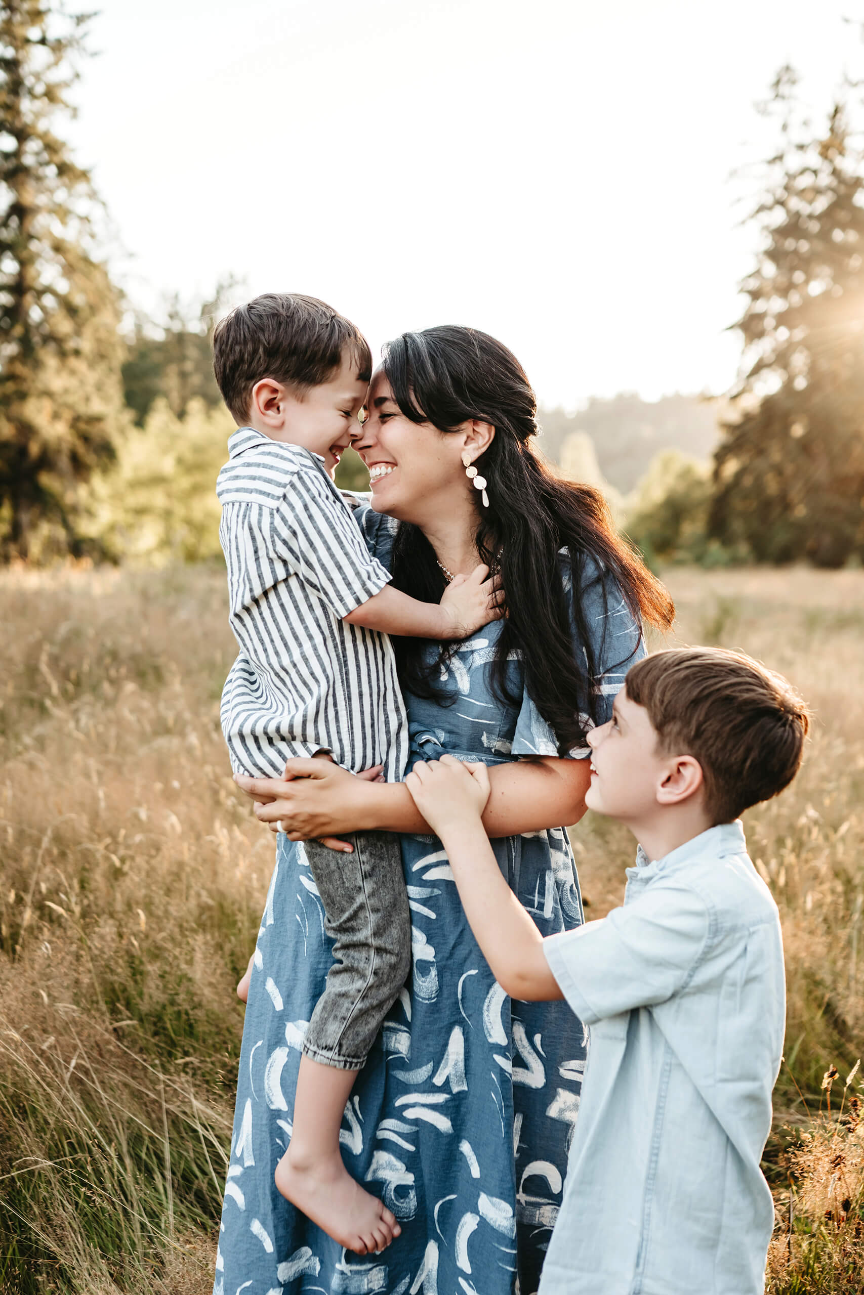 Mother and younger son touching noses and smiling, older brother holding mom’s arm in a grassy field.