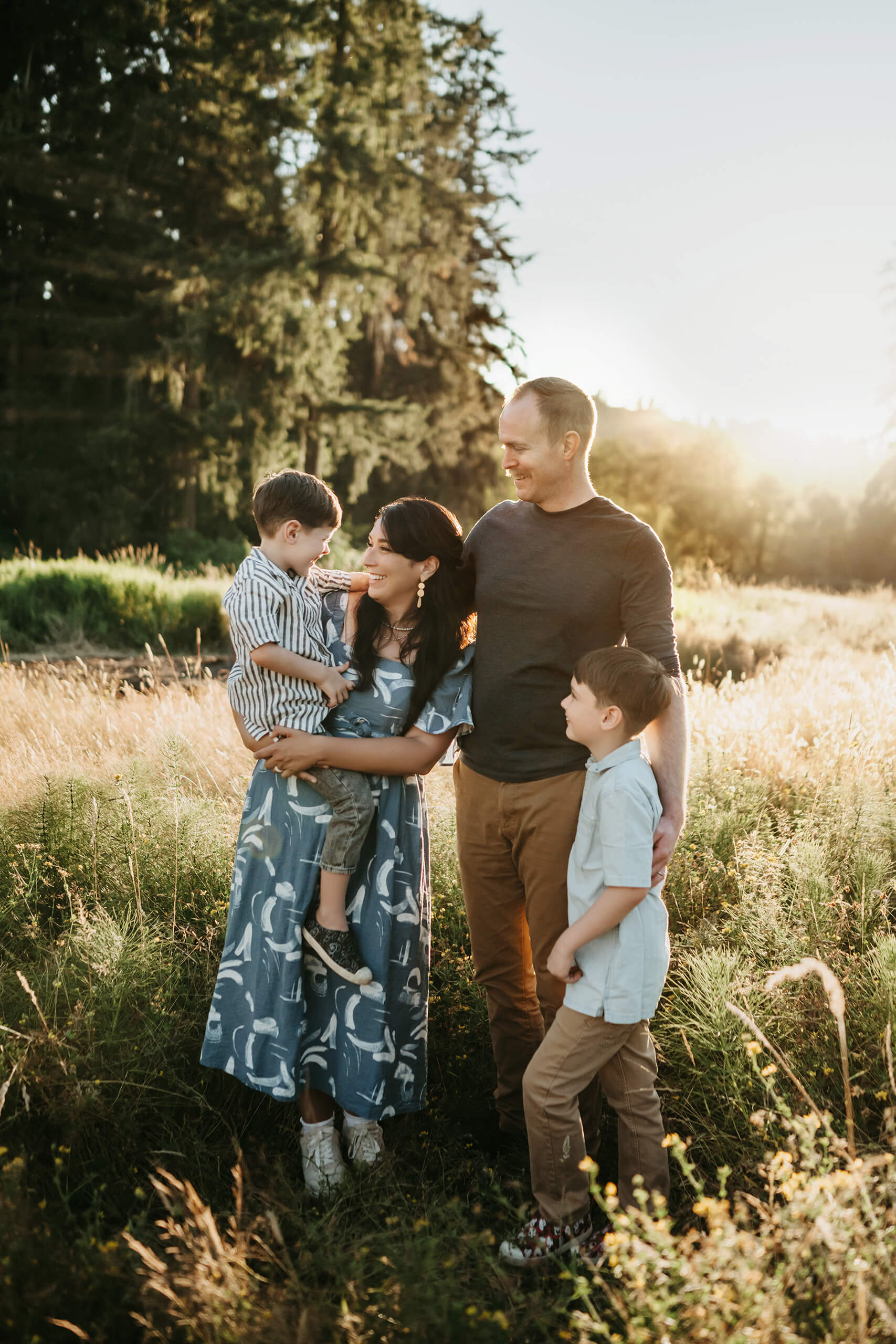 Parents and two boys standing in tall grass at golden hour, mom holding younger son as everyone shares a relaxed smile.