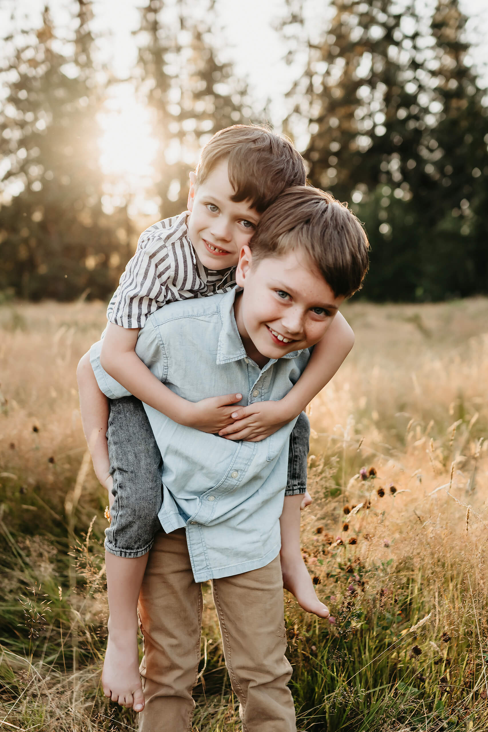 Older brother giving younger brother a piggyback ride, both grinning with sunset flaring through background trees.
