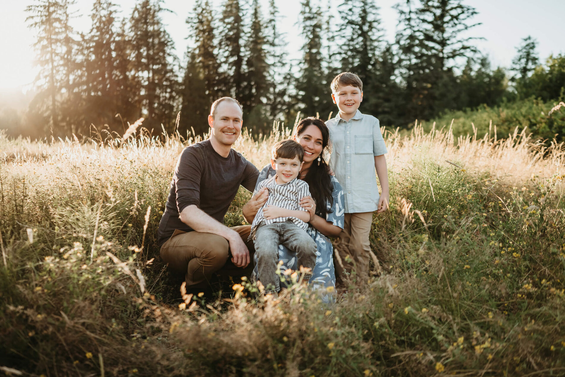 Family of four sitting and kneeling together in a grassy field, sunlight streaming through trees for a warm, candid portrait.