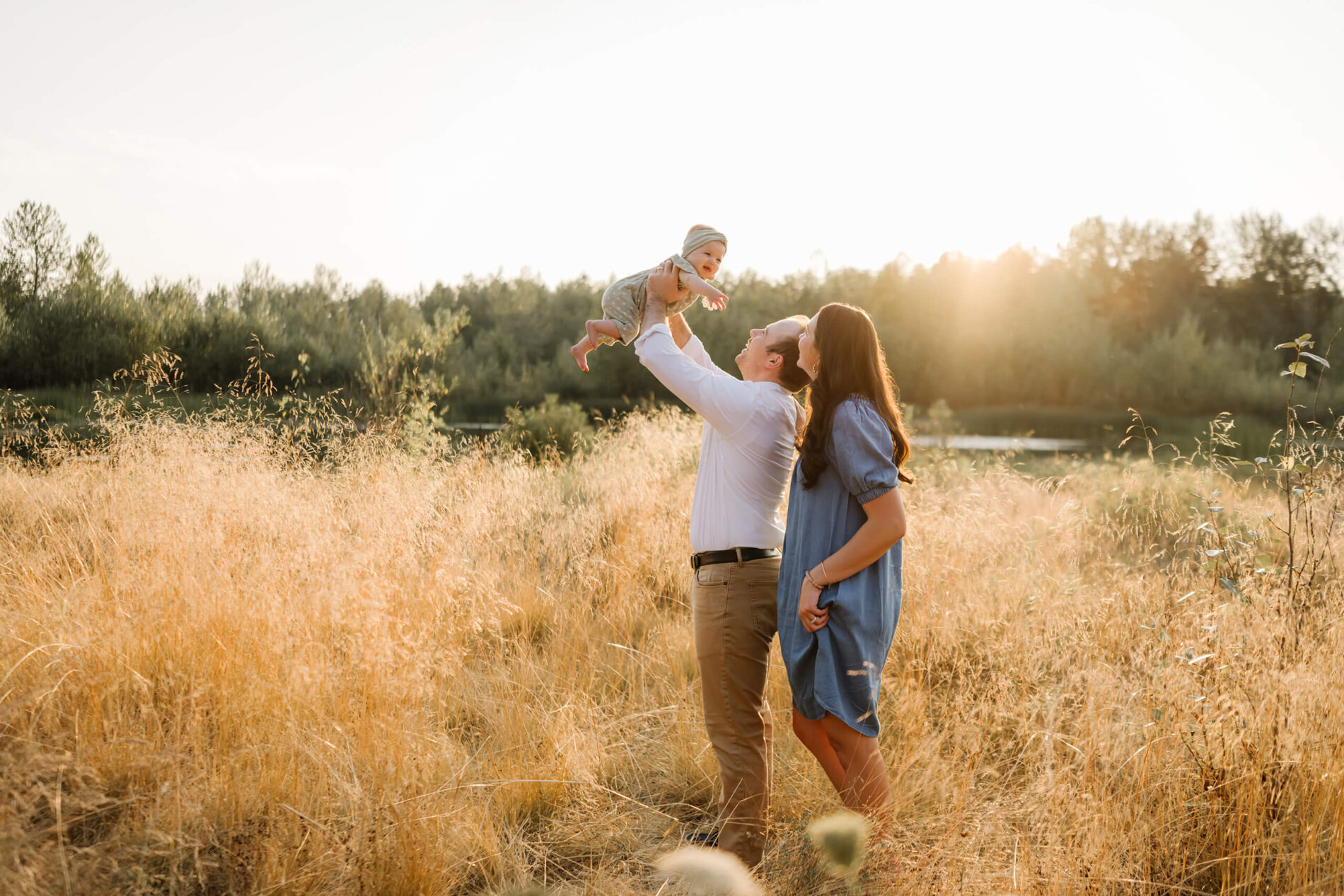 Golden-hour family photo in meadow near Bellevue: dad lifts smiling baby sky-high, mom in blue dress watches, honey-gold grass with pond and trees behind.