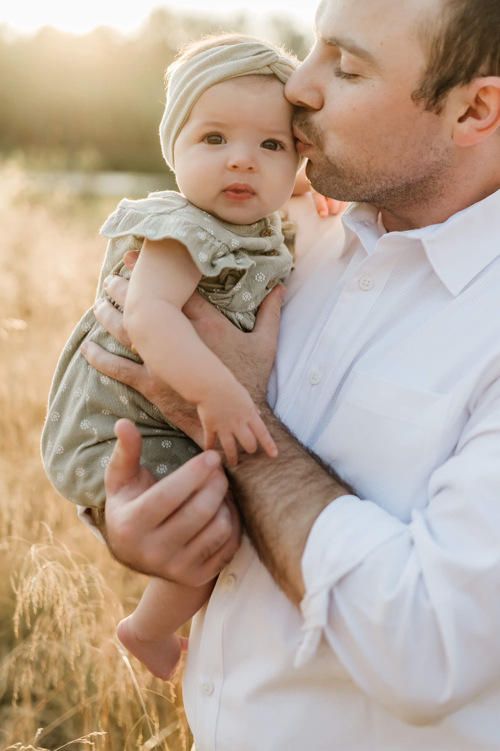 Father kissing baby’s cheek while holding her close in evening light.