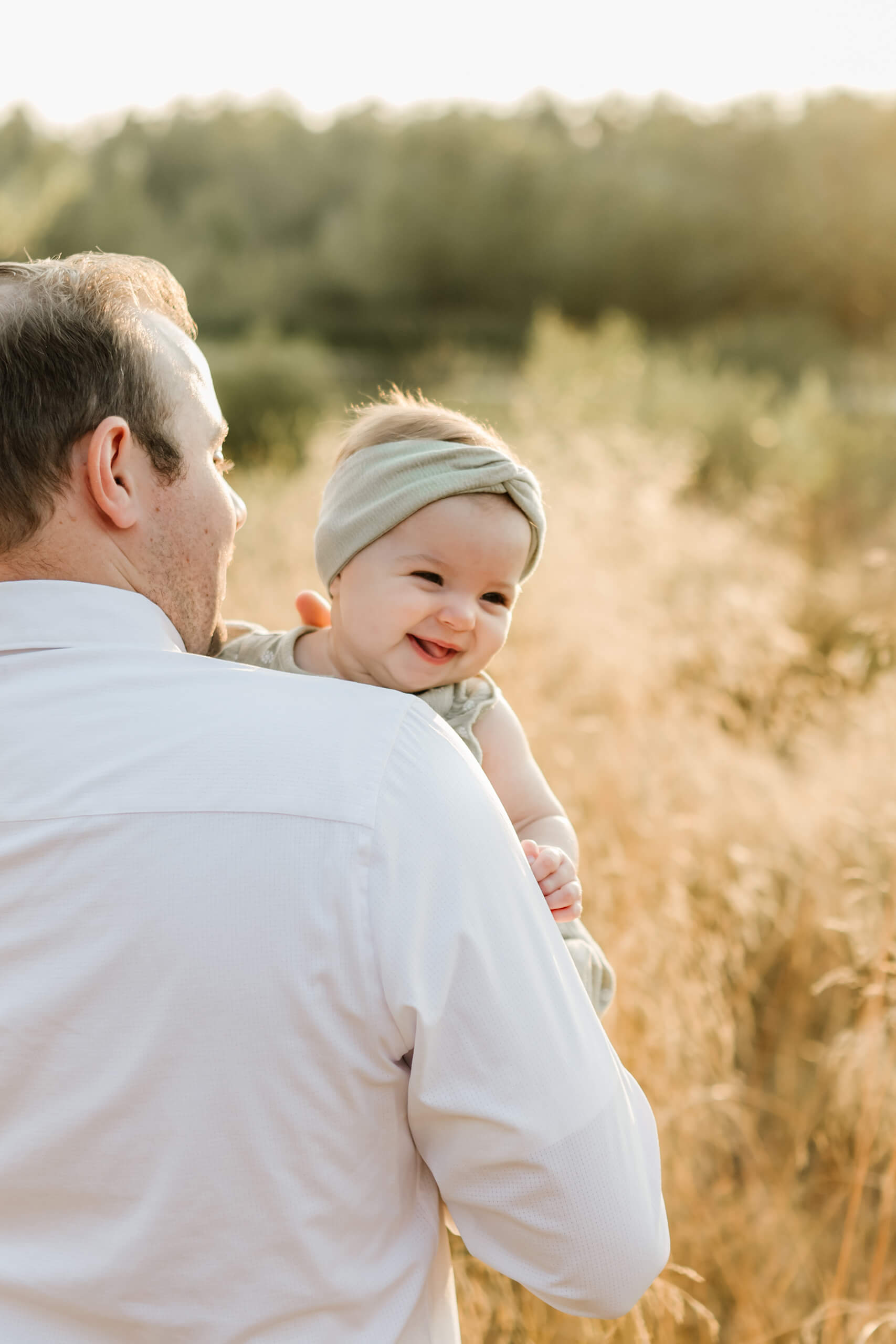 Smiling baby peeking over dad’s shoulder in sun-lit tall grass.