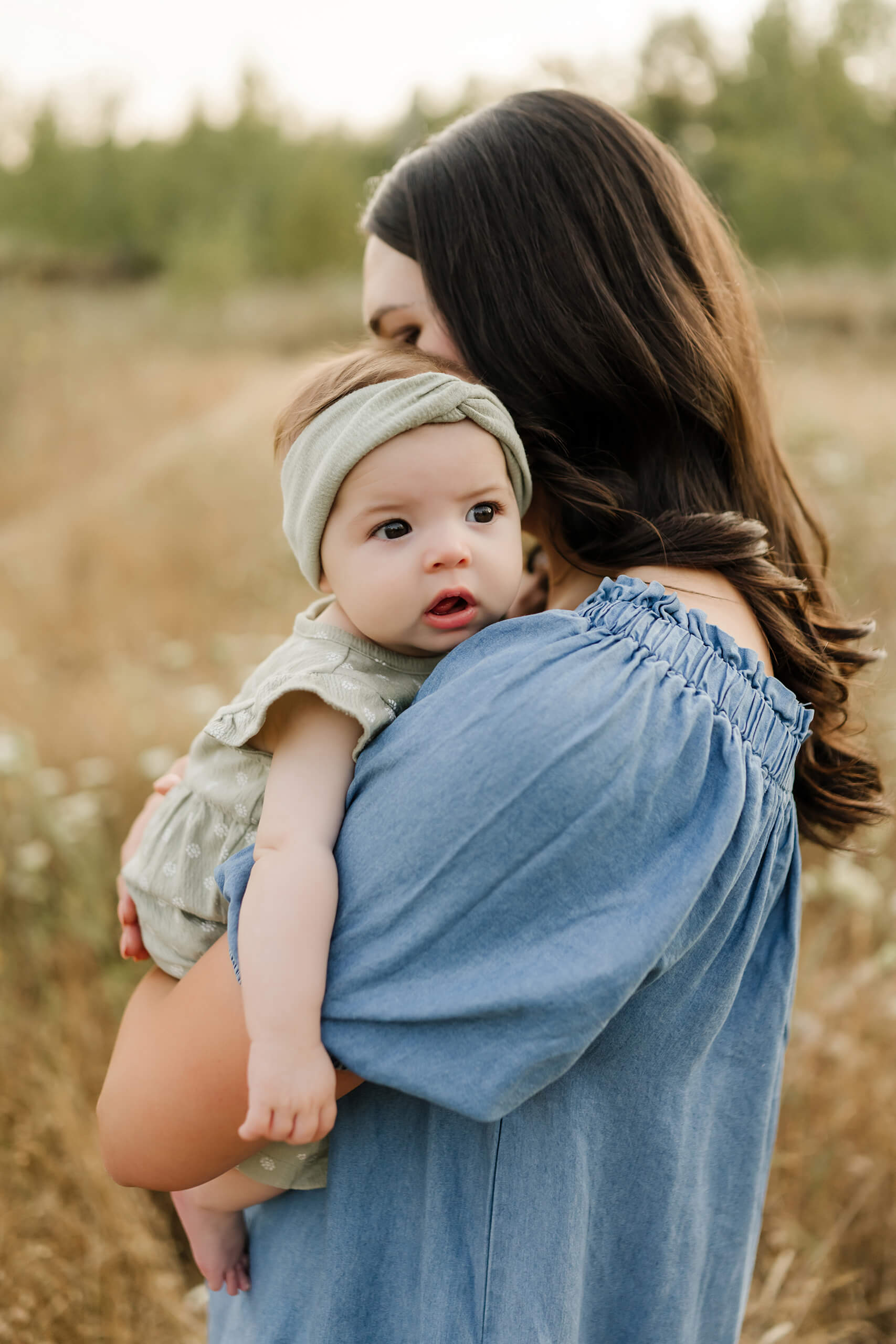 Baby looking over mom’s shoulder with soft evening backdrop.