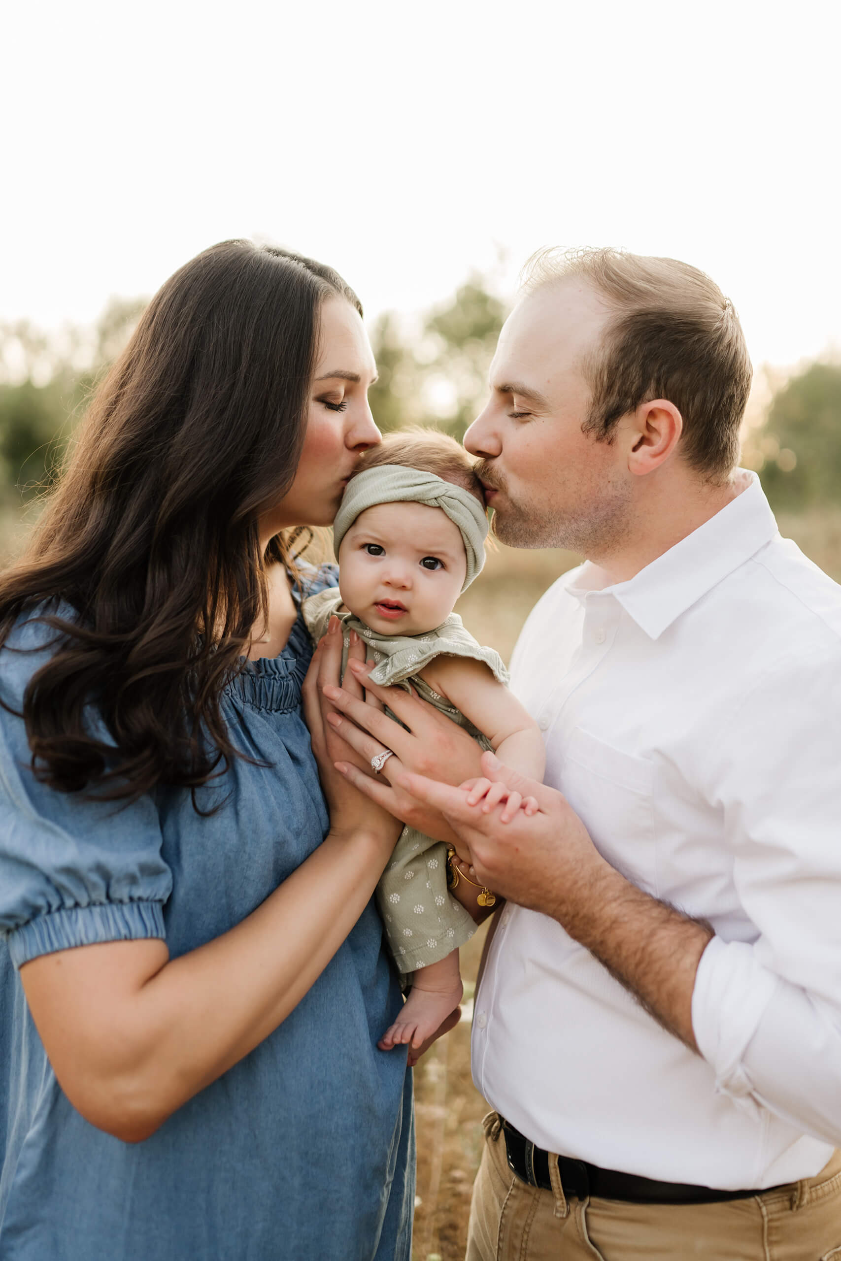 Mom and dad both kissing baby’s head in a tender family moment at dusk.