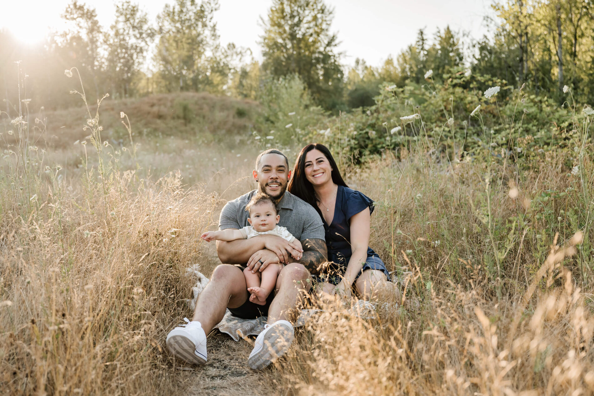 Young family sitting on a blanket in golden summer grass at Redmond park—dad cradles baby on his lap while mom smiles beside them in warm evening light.