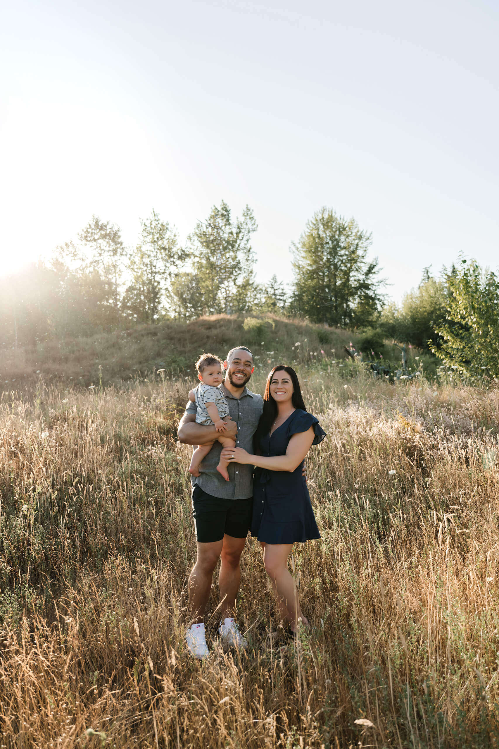 Family of three standing together in a hillside meadow at golden hour, dad cradling their toddler.