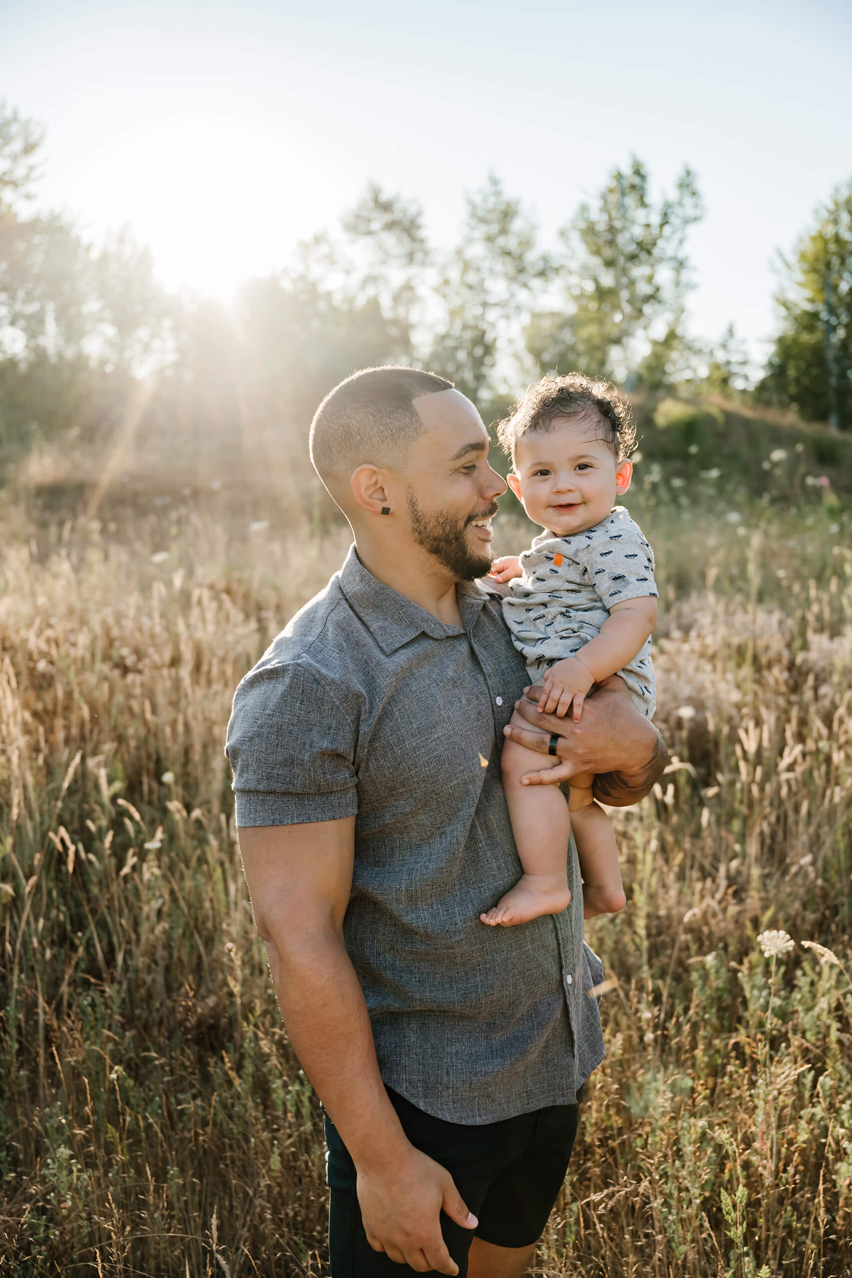 Father gazing at and holding his smiling baby boy against a backdrop of sun-drenched wild grass.