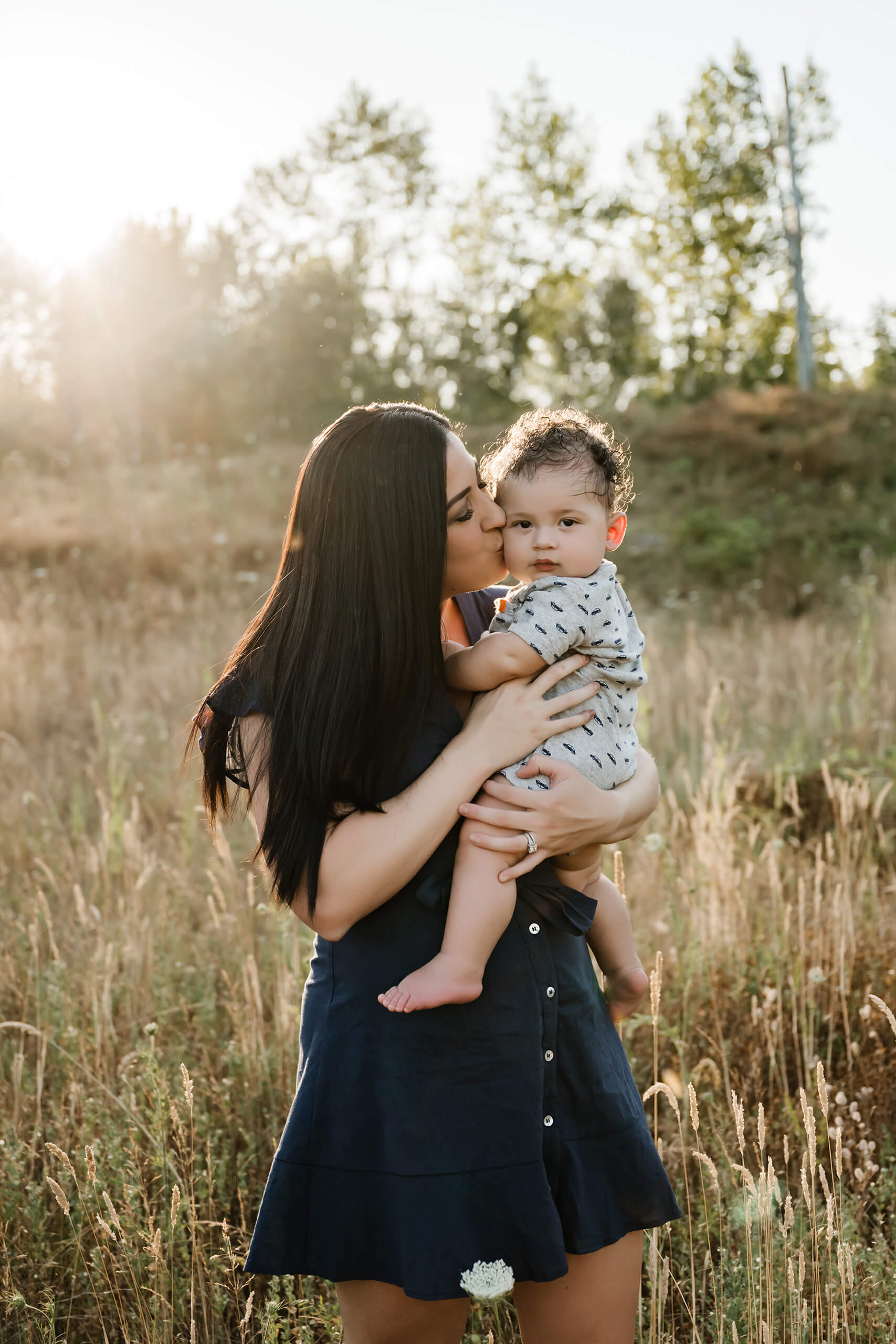 Mother in a navy dress kissing her baby’s cheek while holding him in tall golden grass at sunset.