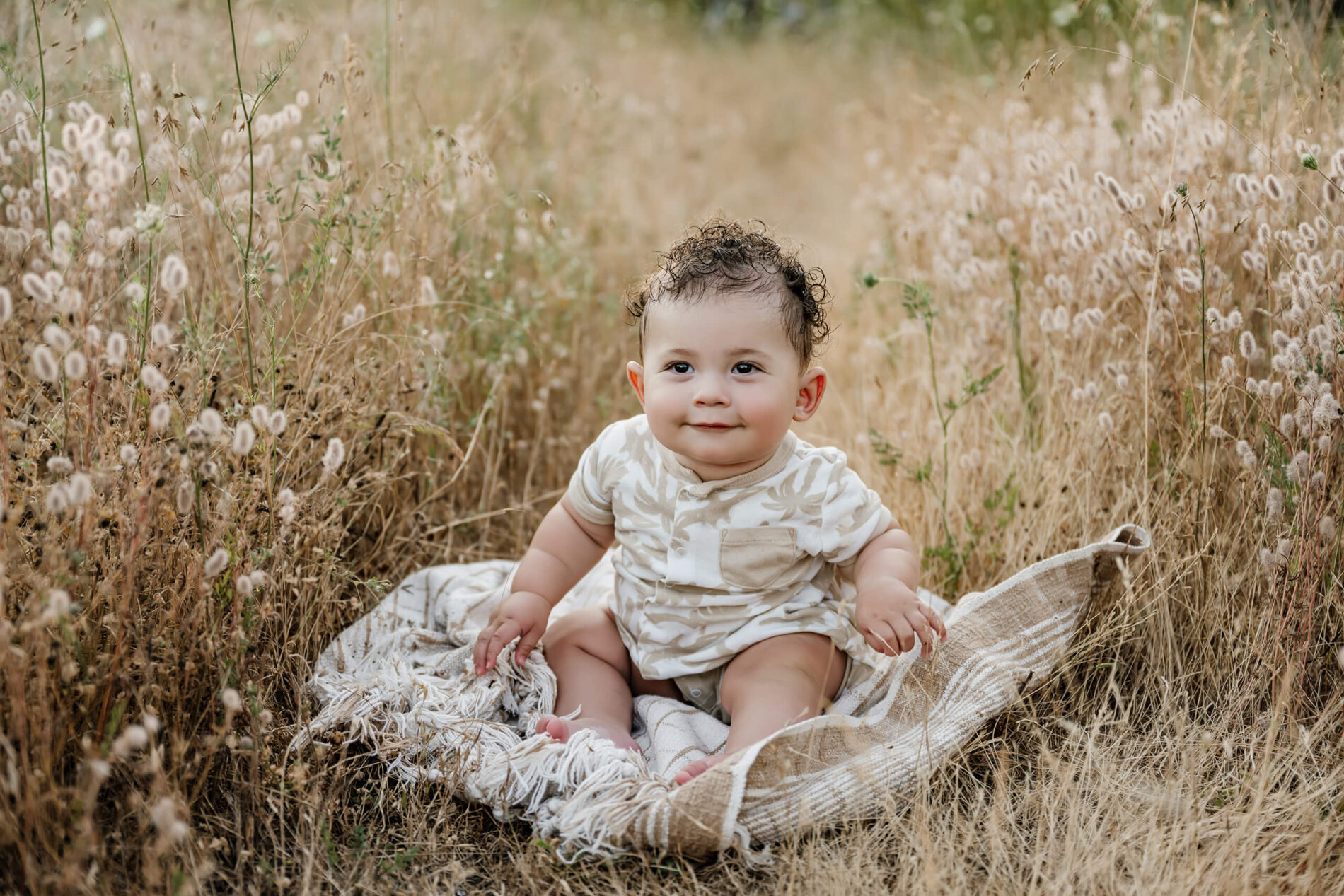 Baby sitting on a striped blanket in a dry summer meadow, surrounded by fluffy tan grasses.
