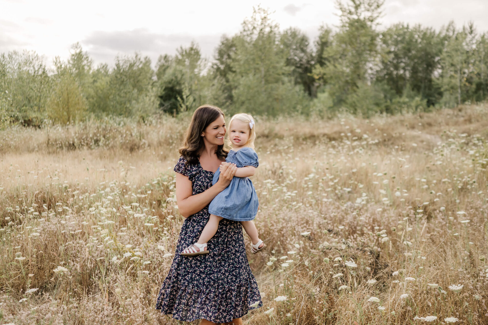 Mother holding toddler daughter in denim dress while standing in a wildflower-filled meadow at Redmond park.