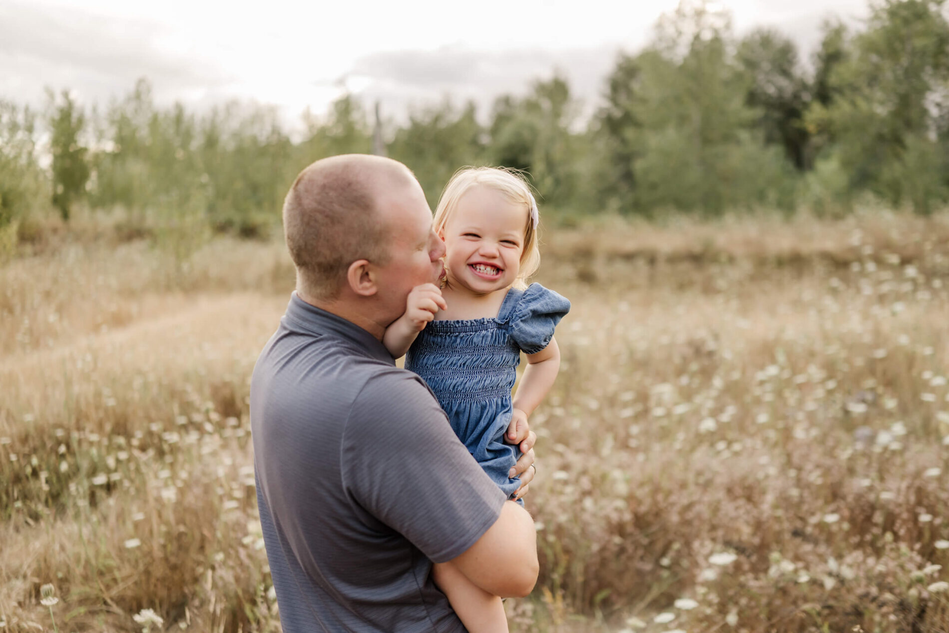 Dad holding his laughing toddler daughter in a wildflower meadow in Redmond while giving her a playful kiss on the cheek.