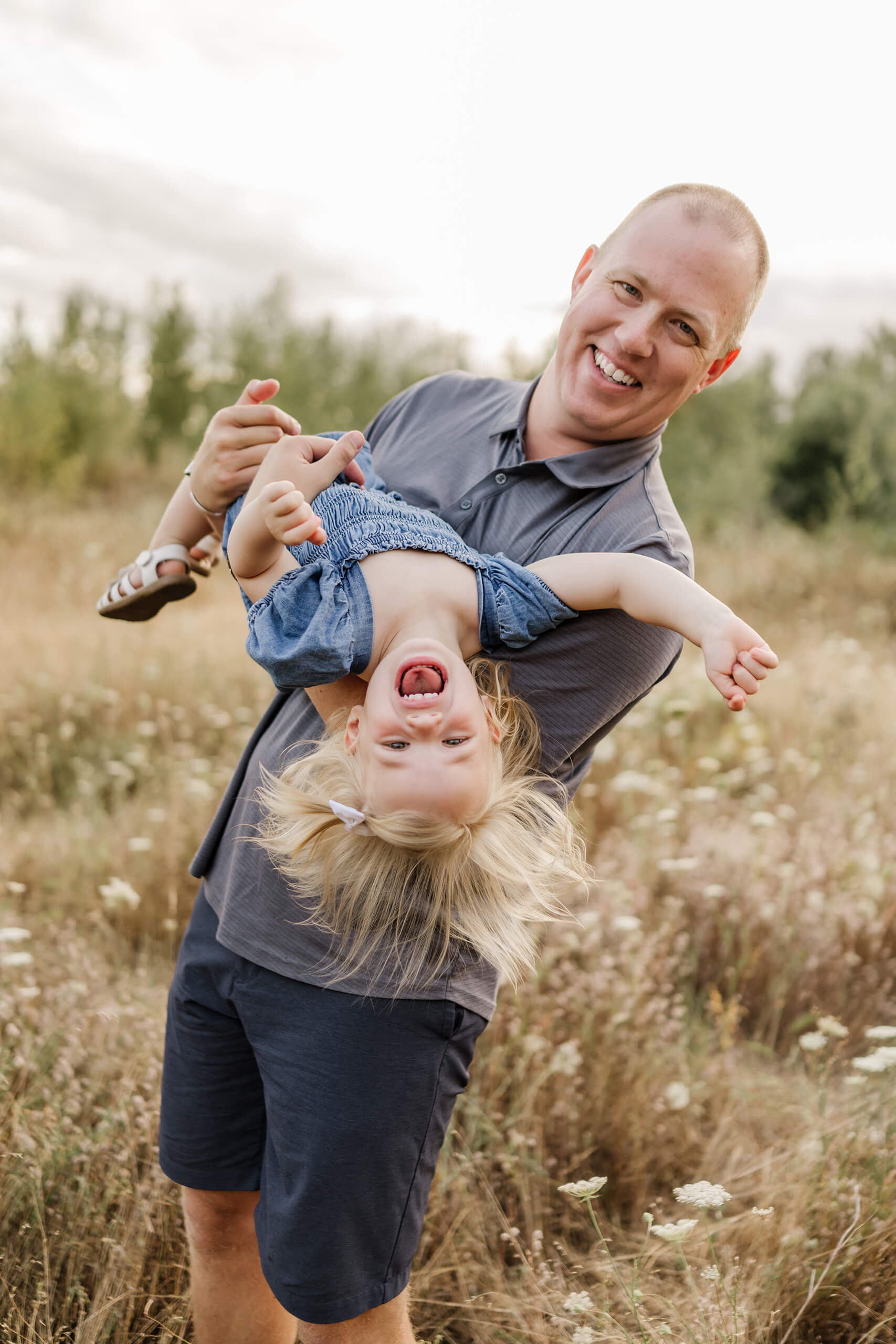 Father swinging daughter upside-down; she laughs with mouth wide open against a backdrop of tall grass.