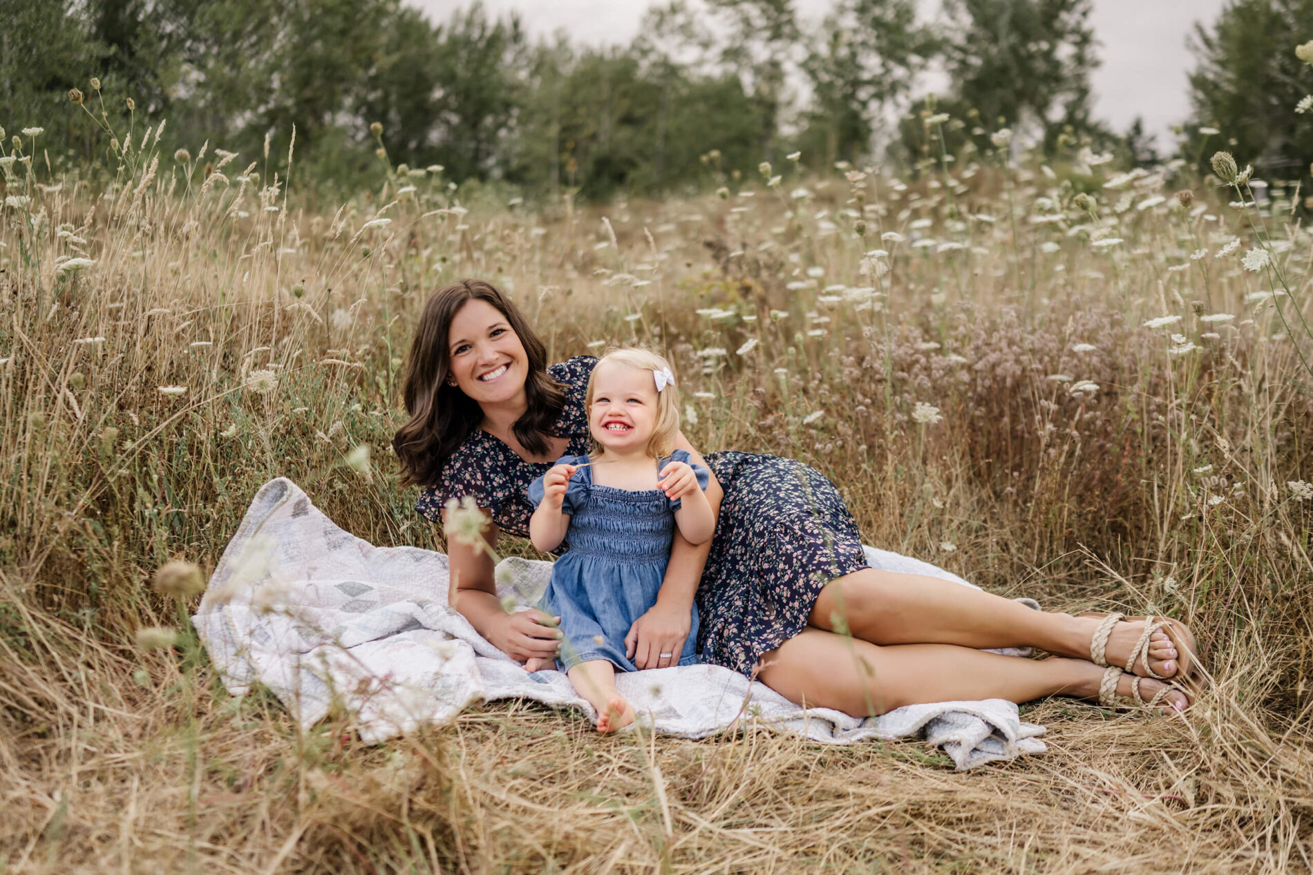 Mom and daughter relaxing on a quilt in tall grass, grinning amid blooming wildflowers.