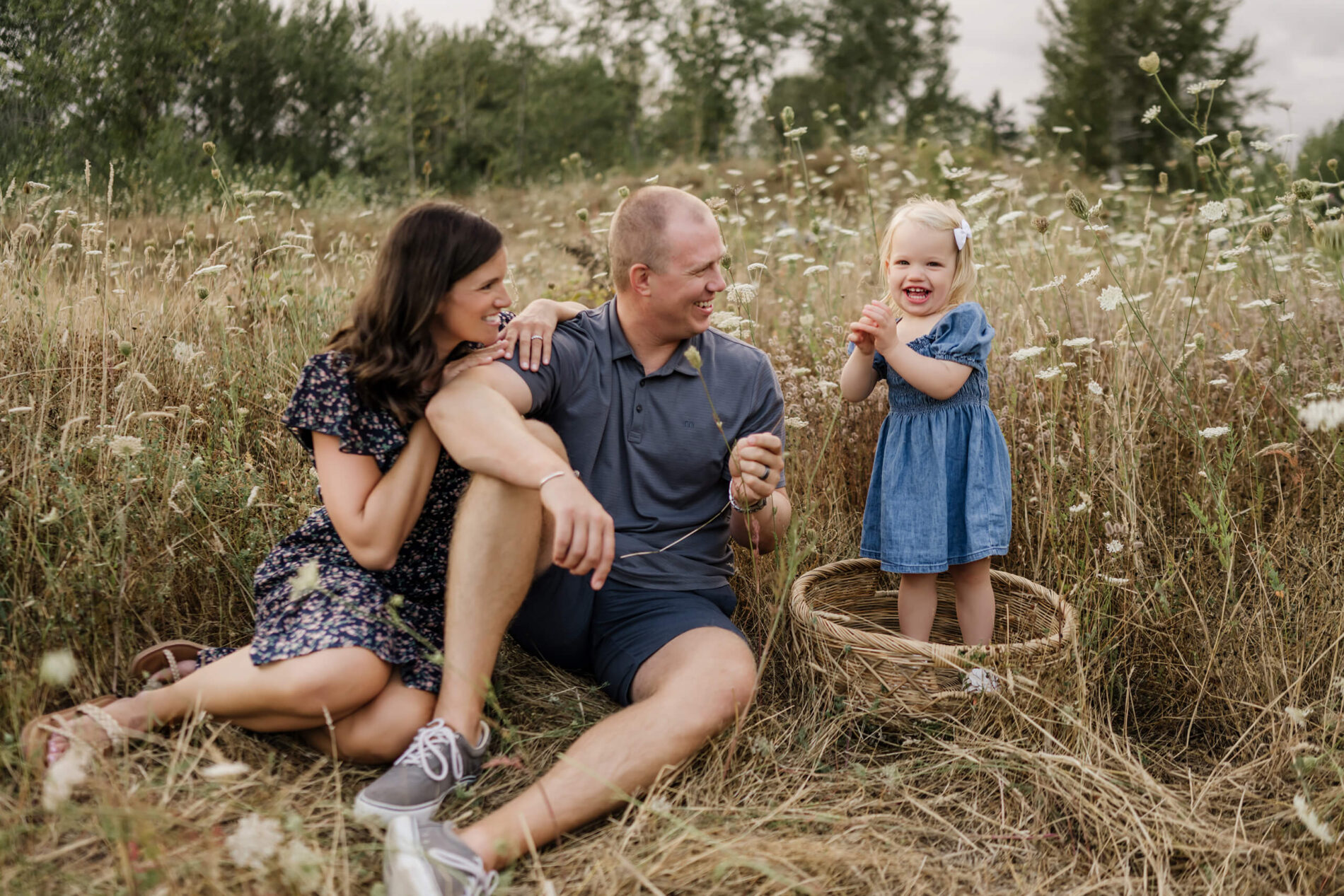 Family sitting among Queen Anne’s lace; daughter stands in a wicker basket, parents laughing beside her.
