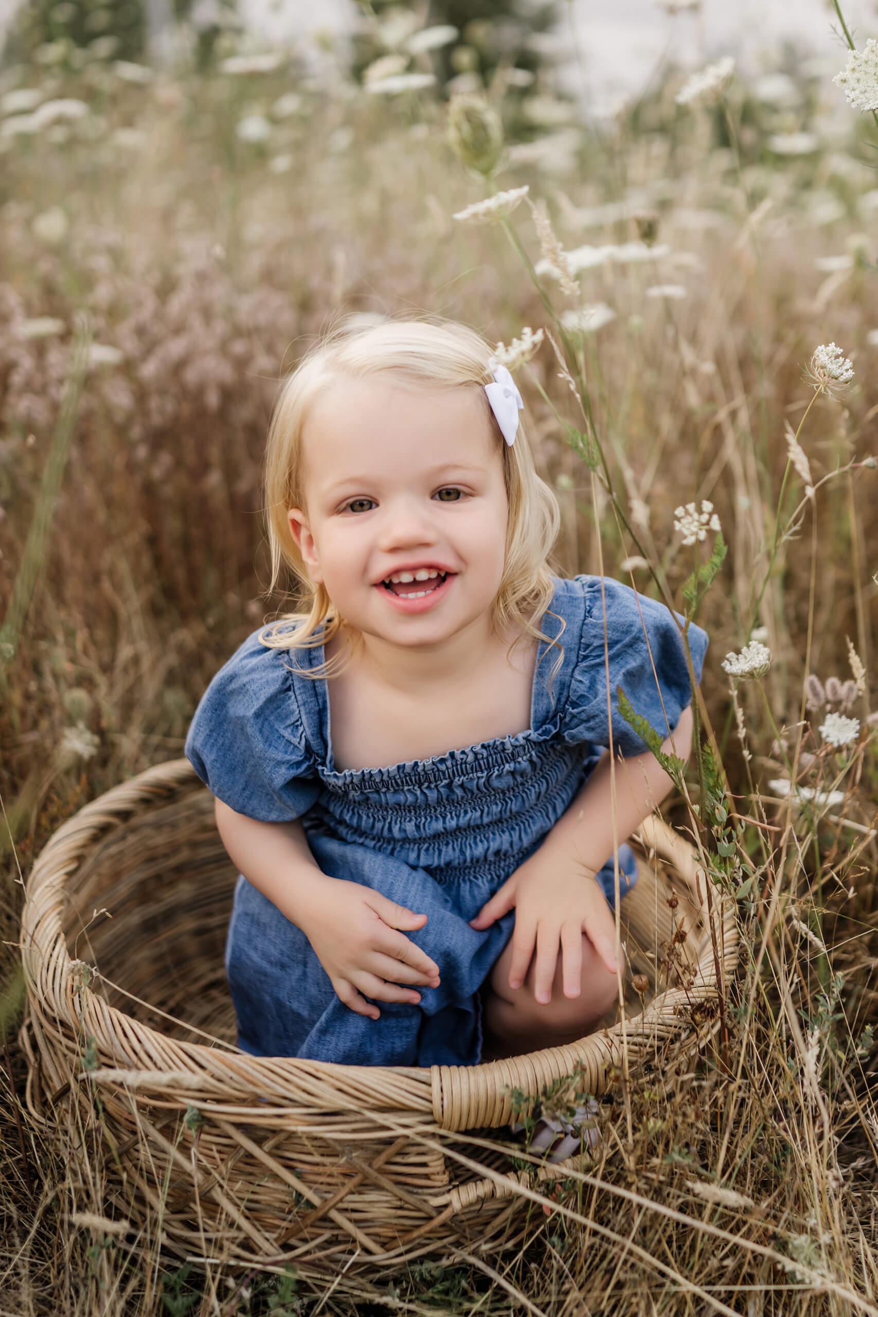 Close-up of happy toddler girl sitting inside a wicker basket, surrounded by delicate wildflowers.