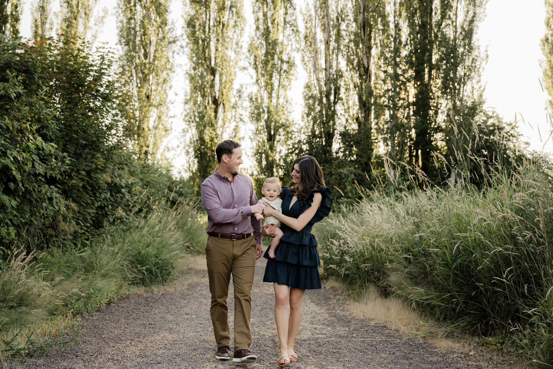 During a family photoshoot, parents strolling down a tree-lined path, smiling at each other while holding their baby between them.