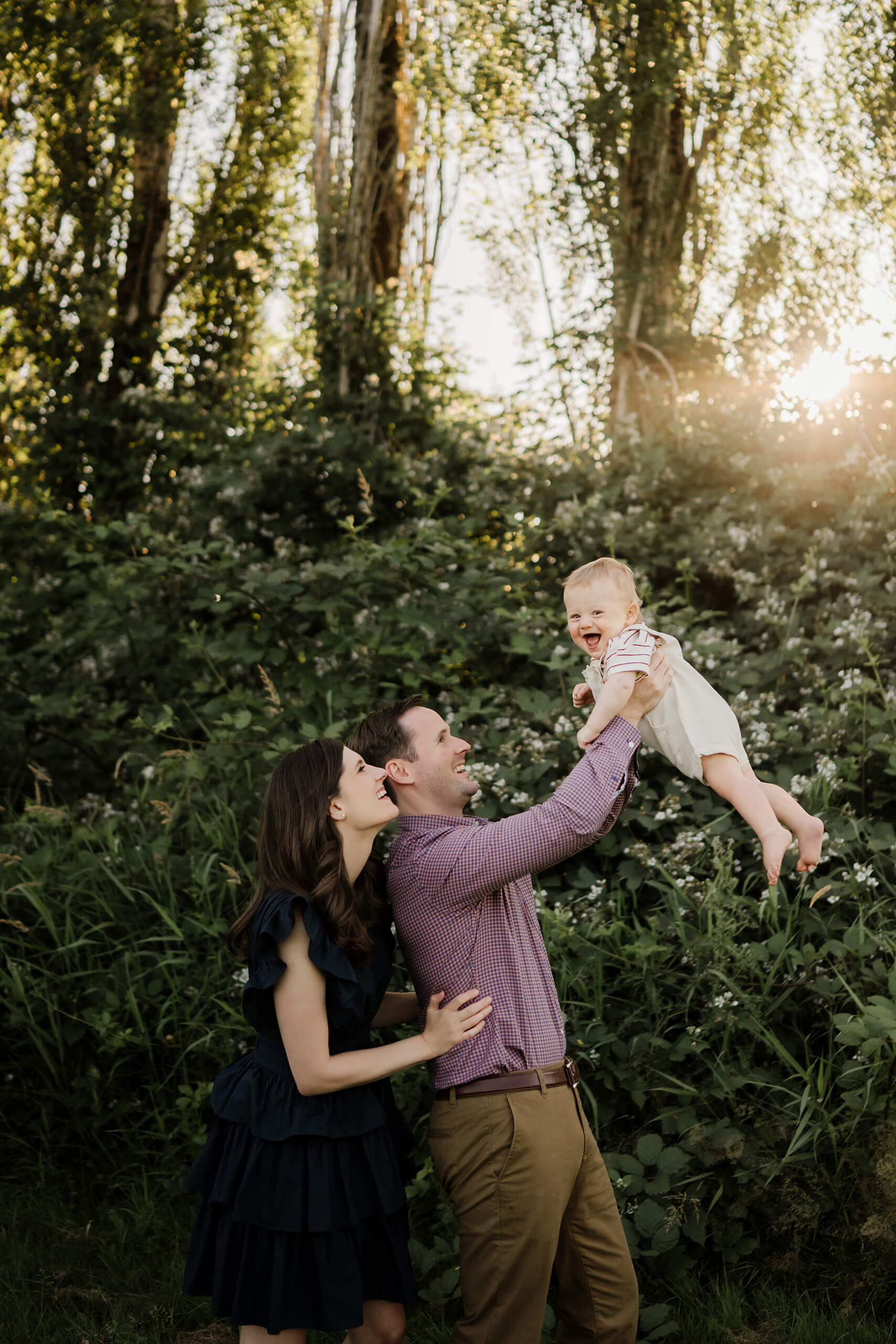 Dad hoists laughing baby into the air as mom looks on, evening sun filtering through leafy greenery.