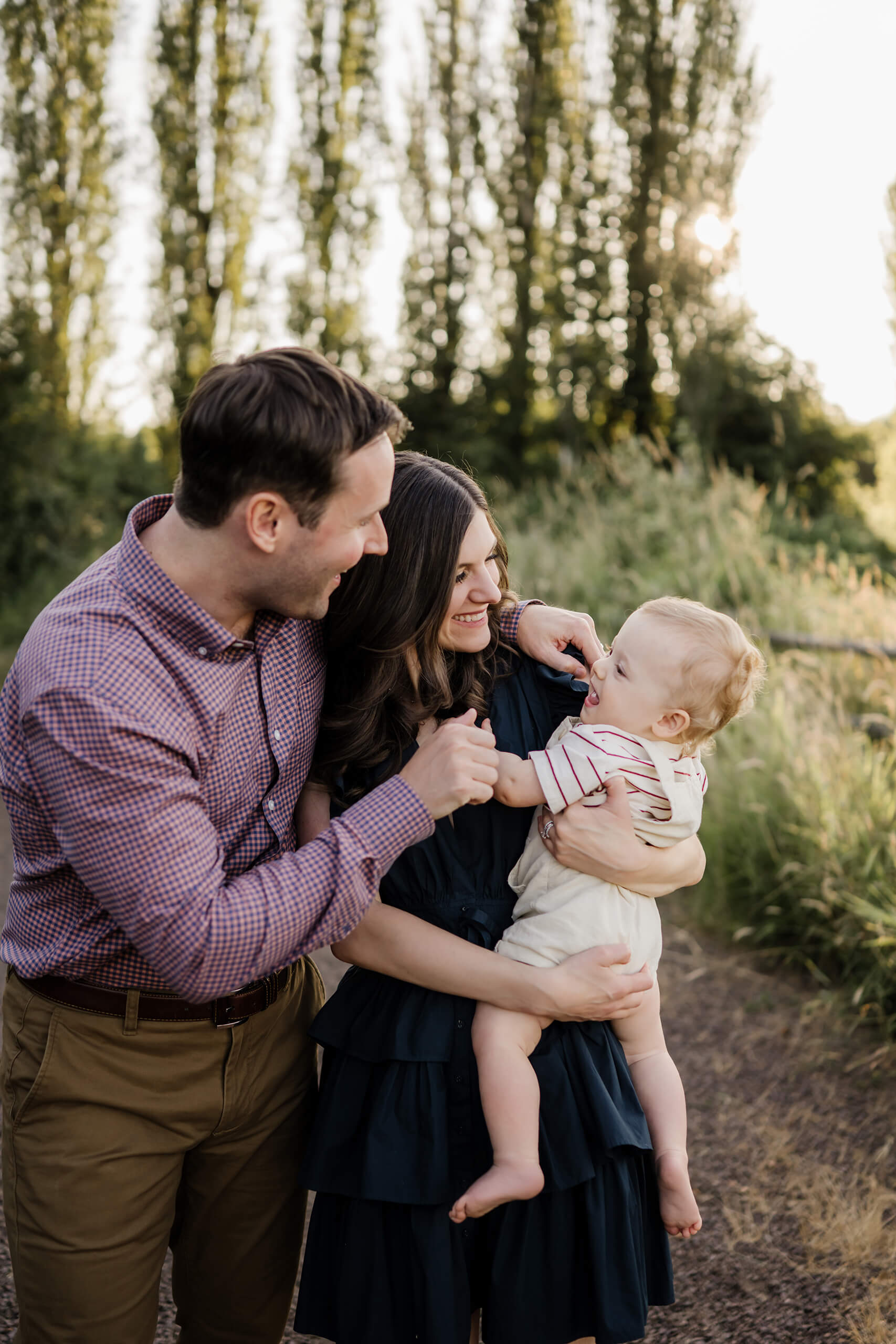 Parents tickling their baby in mom’s arms, all three laughing on a wooded trail.