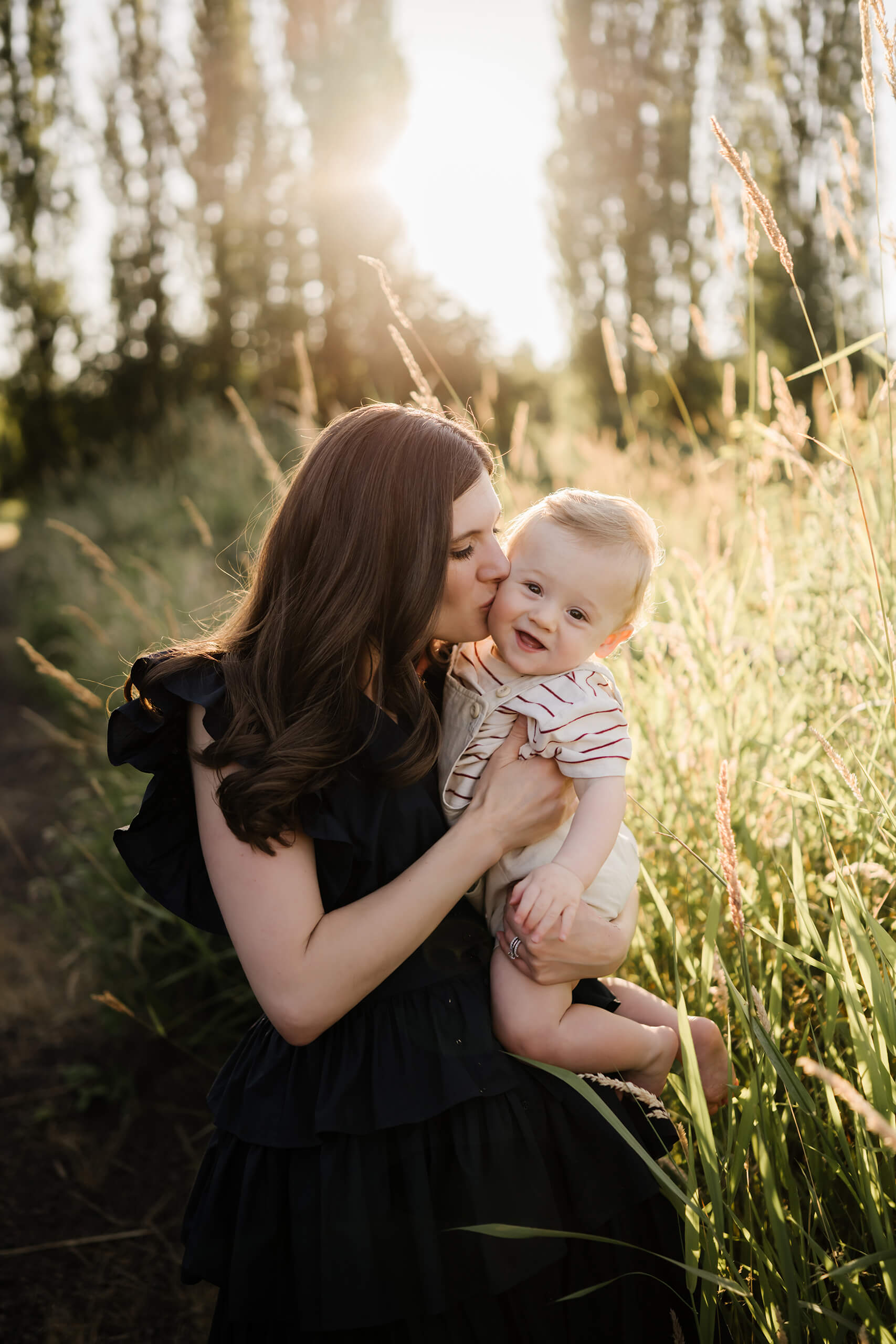 Mom kissing her baby’s cheek in tall back-lit grass, golden sun glowing behind.