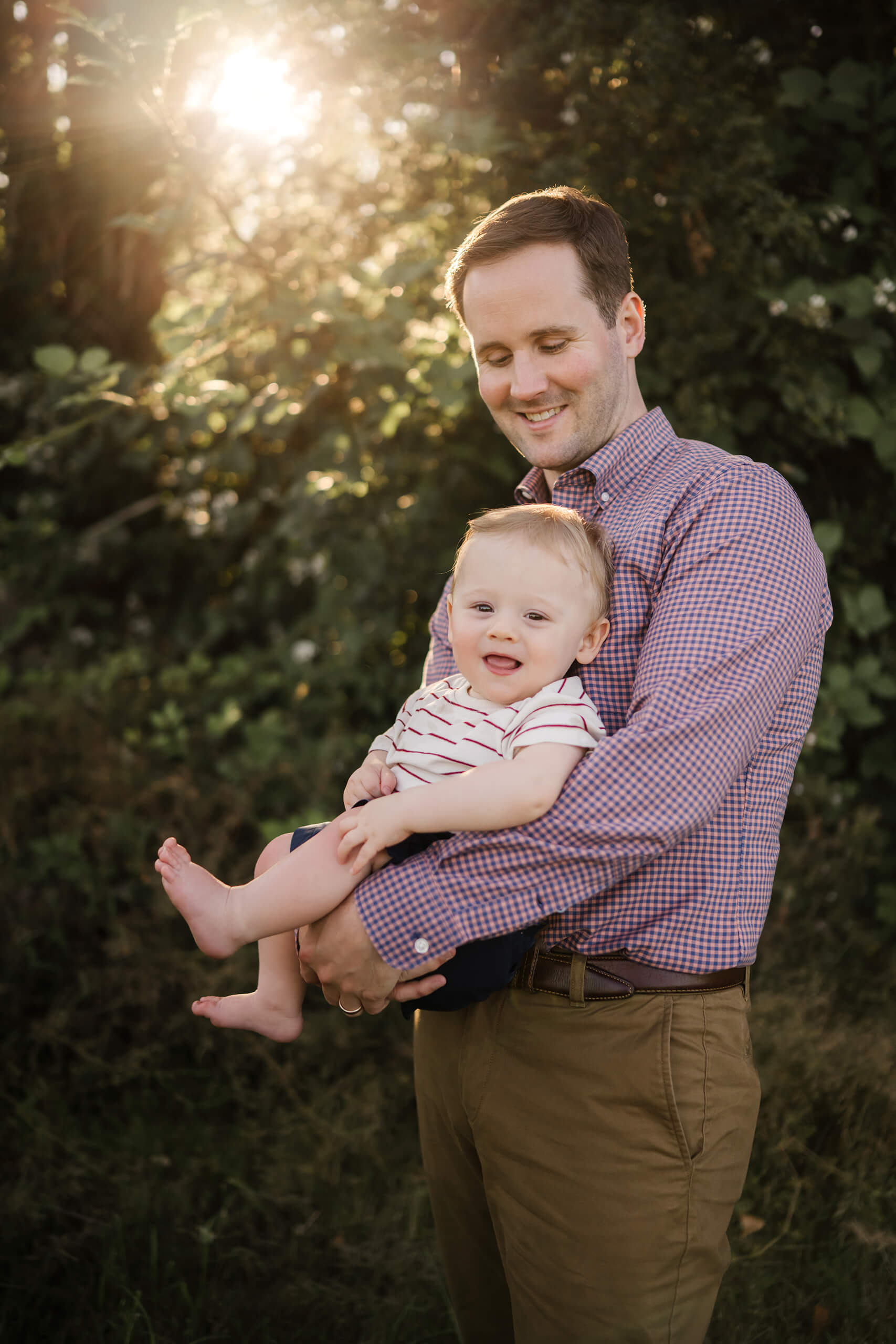 Dad cradling smiling baby in warm sunset light with leafy background.