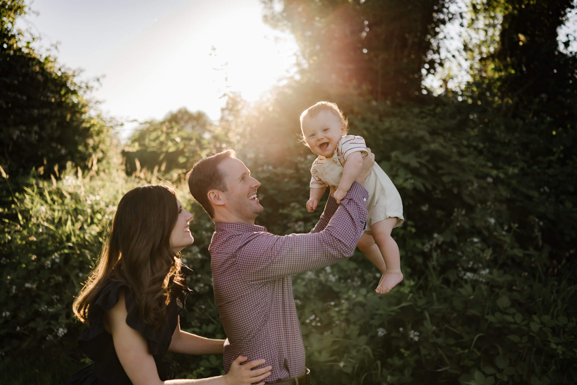 Father lifting cheerful baby toward the light while mom steadies him, sun flare highlighting the moment.