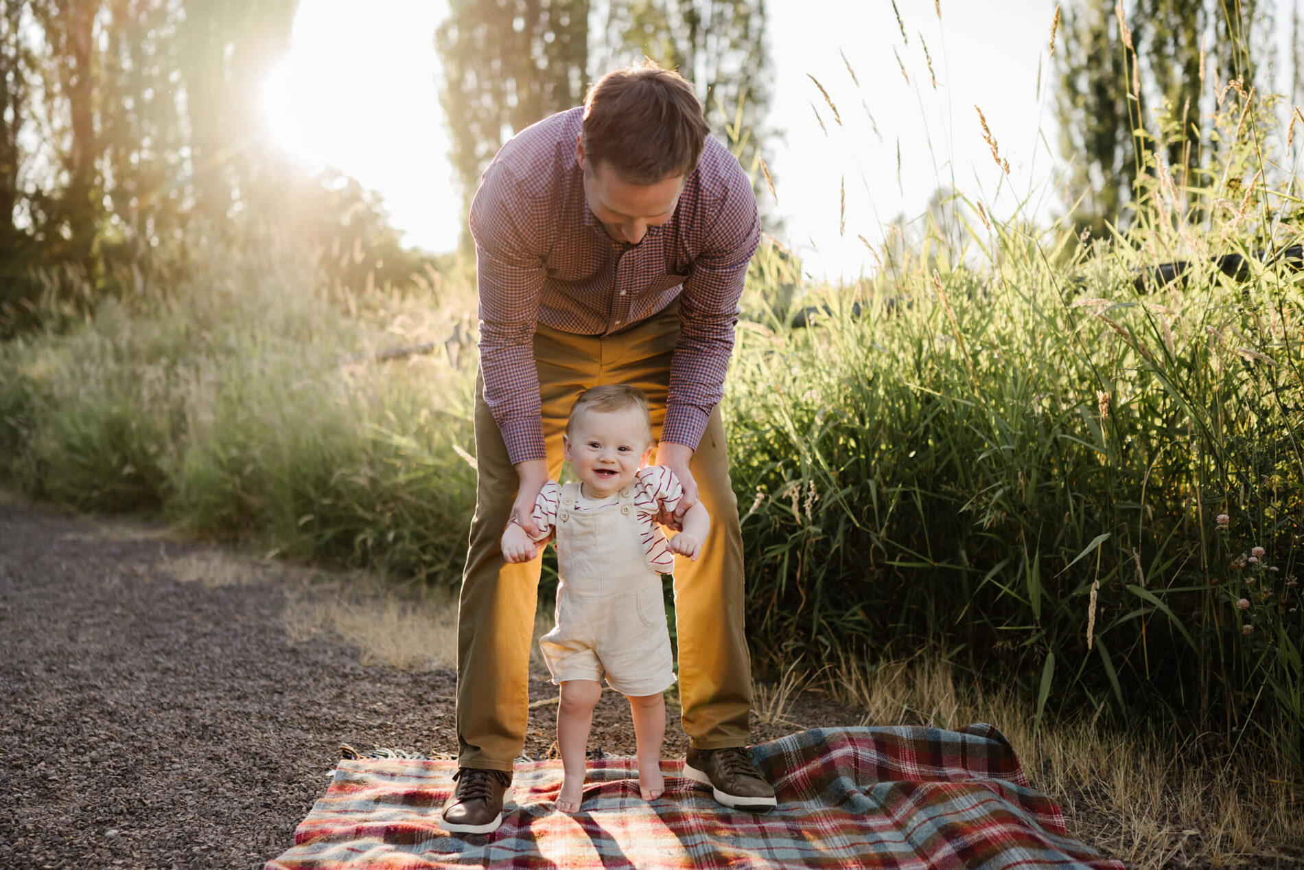 Back-lit evening scene of dad helping his barefoot baby stand on a plaid picnic blanket.