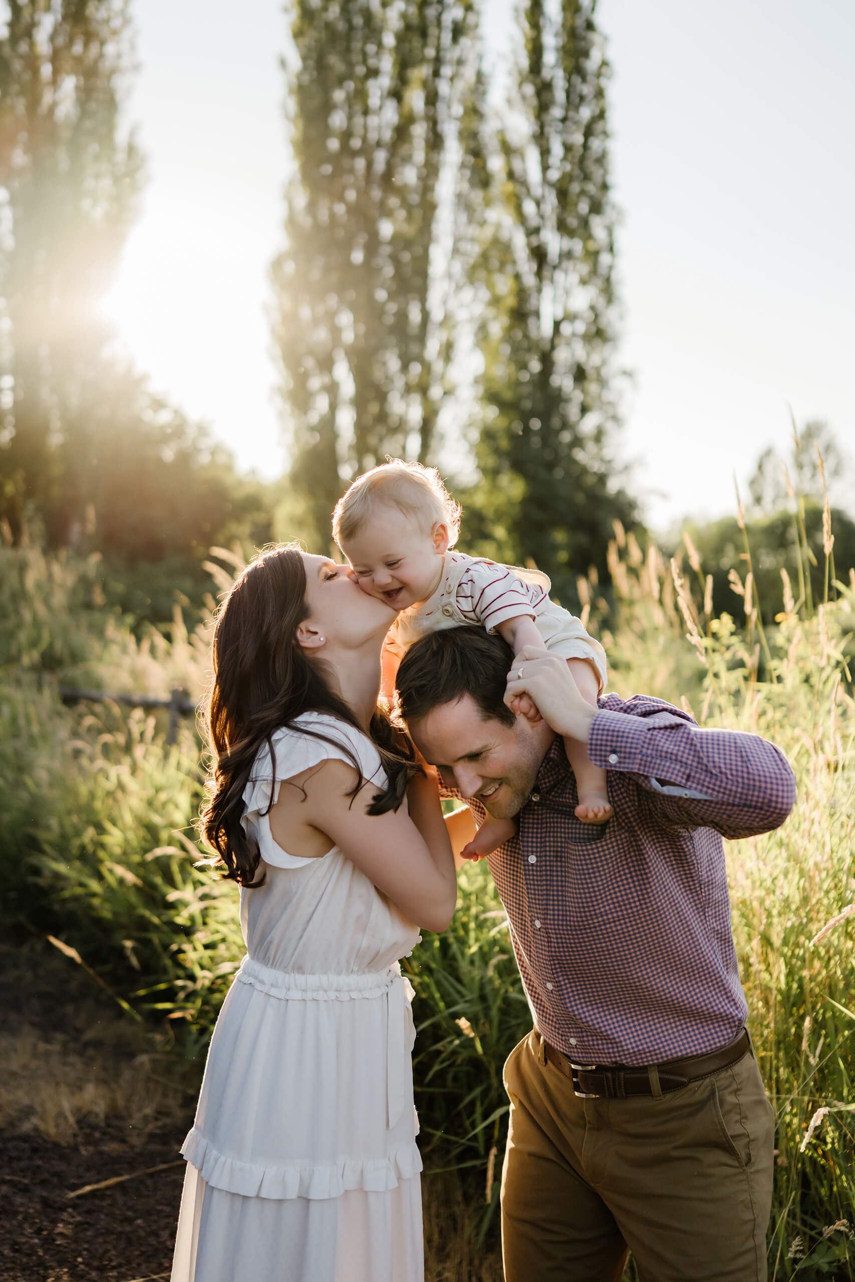 Mom kisses baby sitting on dad’s shoulders as golden light filters through the trees.