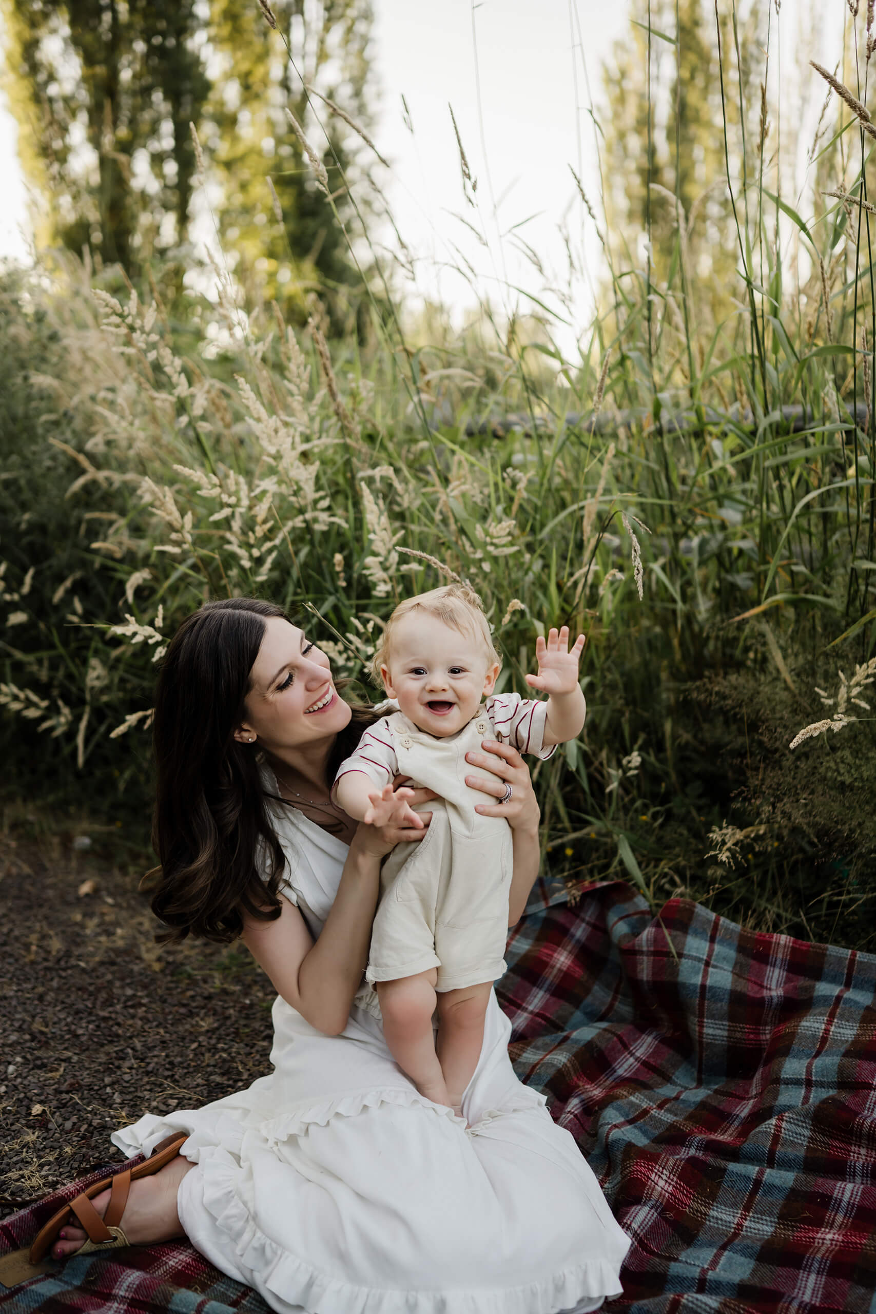 Mom on a blanket lifts her happy baby who waves toward the camera, framed by lush grass.