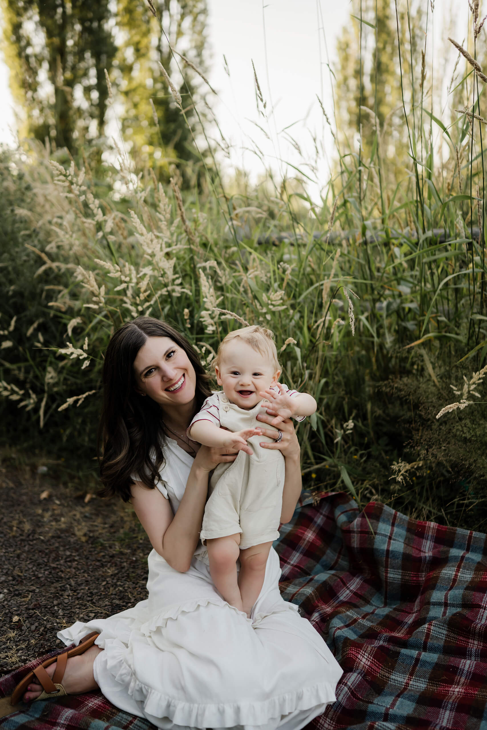 Mom kneels on a blanket, holding a smiling baby upright against a backdrop of wild grass.
